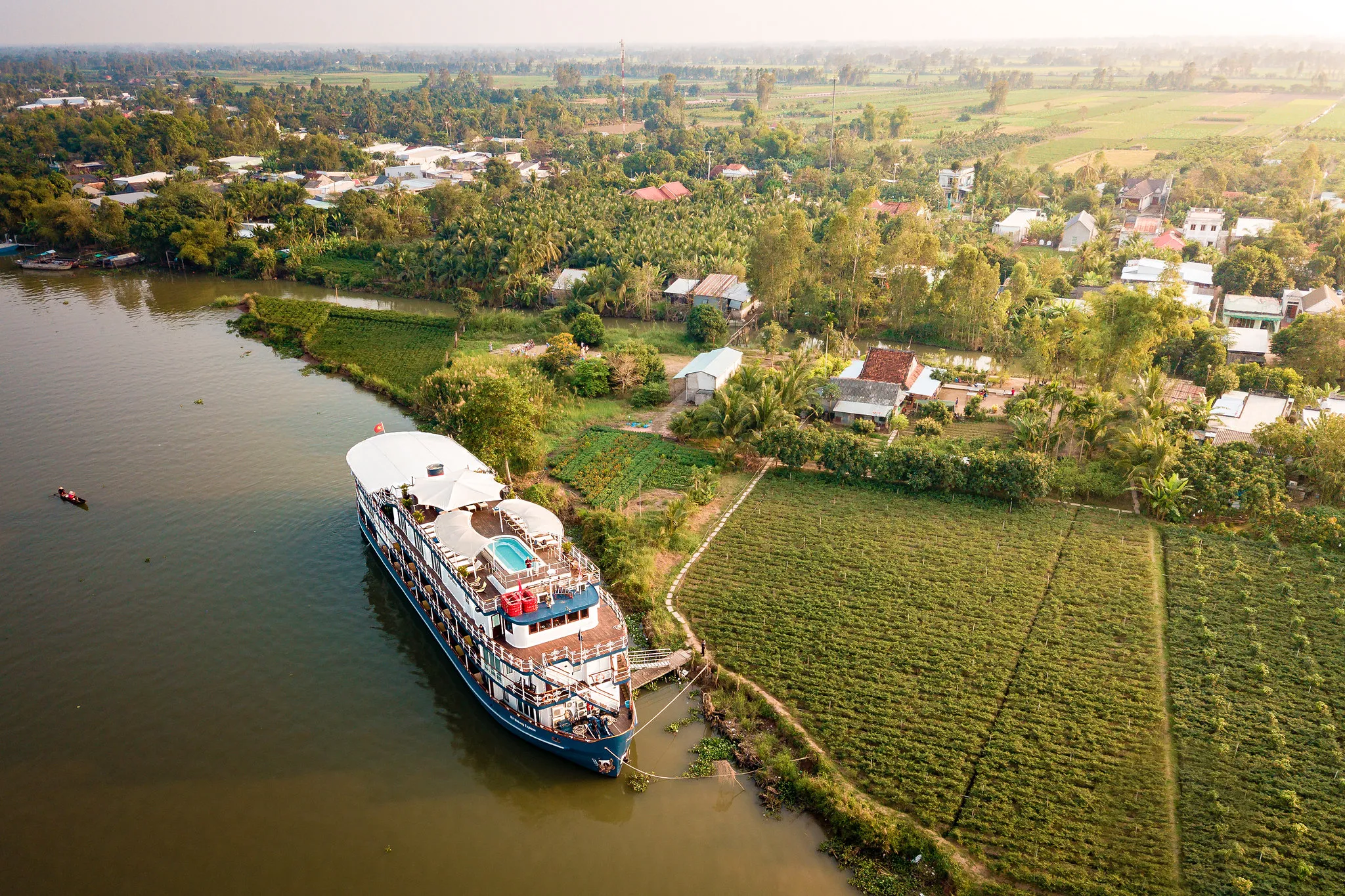 Aerial view of a Heritage Line cruise ship docked along a lush, green riverside village in Asia, surrounded by fields and scattered houses.