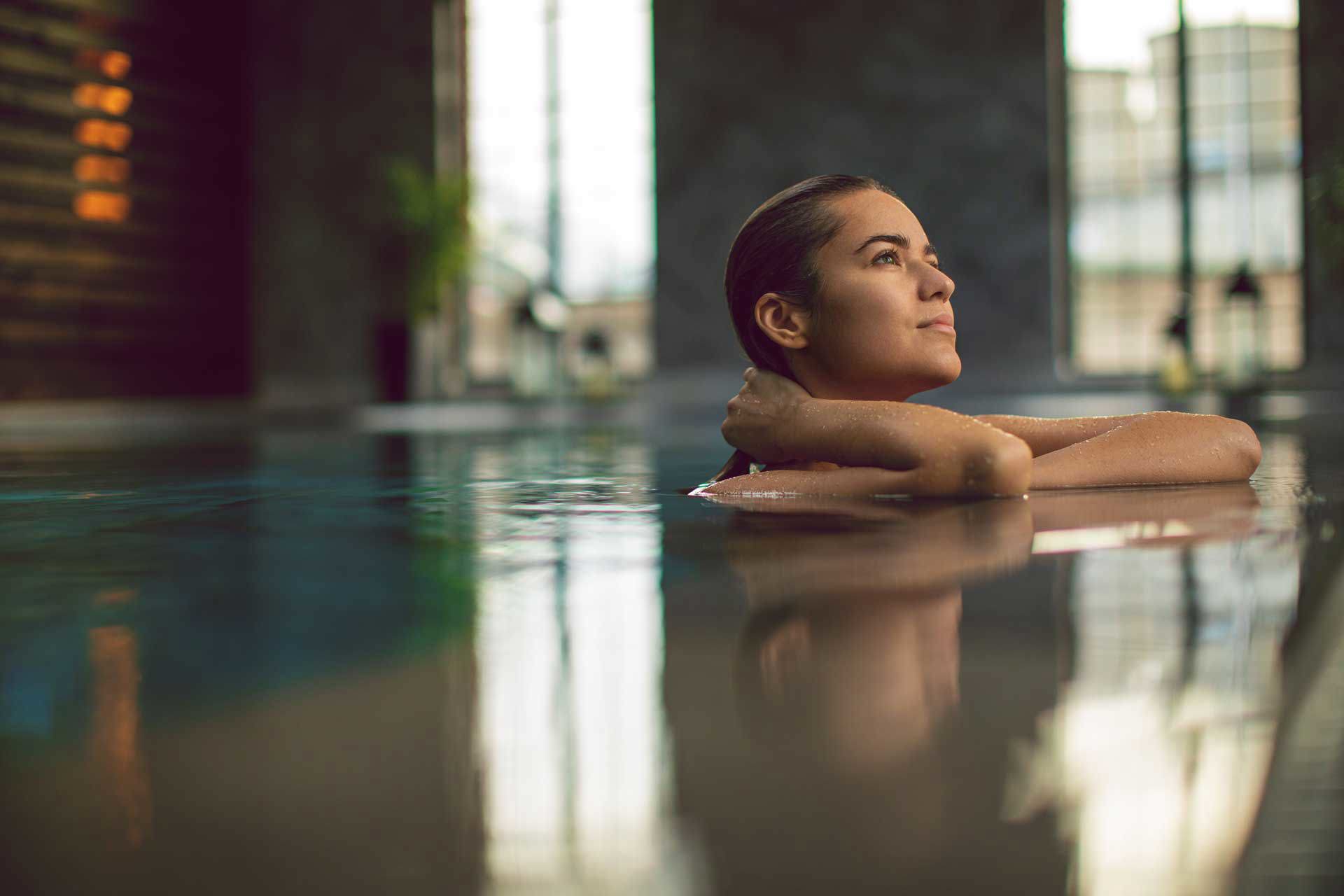 Woman relaxing in a calm indoor pool, resting on the edge and looking towards soft natural light in a tranquil spa setting.