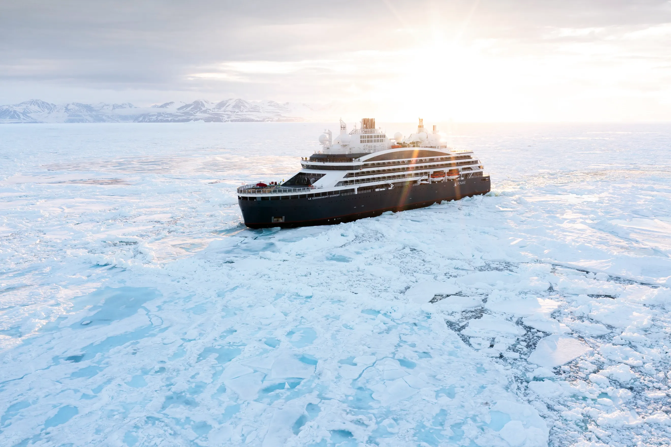 Ponant Explorations expedition ship cruising through icy waters with snow-covered mountains and a low sun on the horizon.