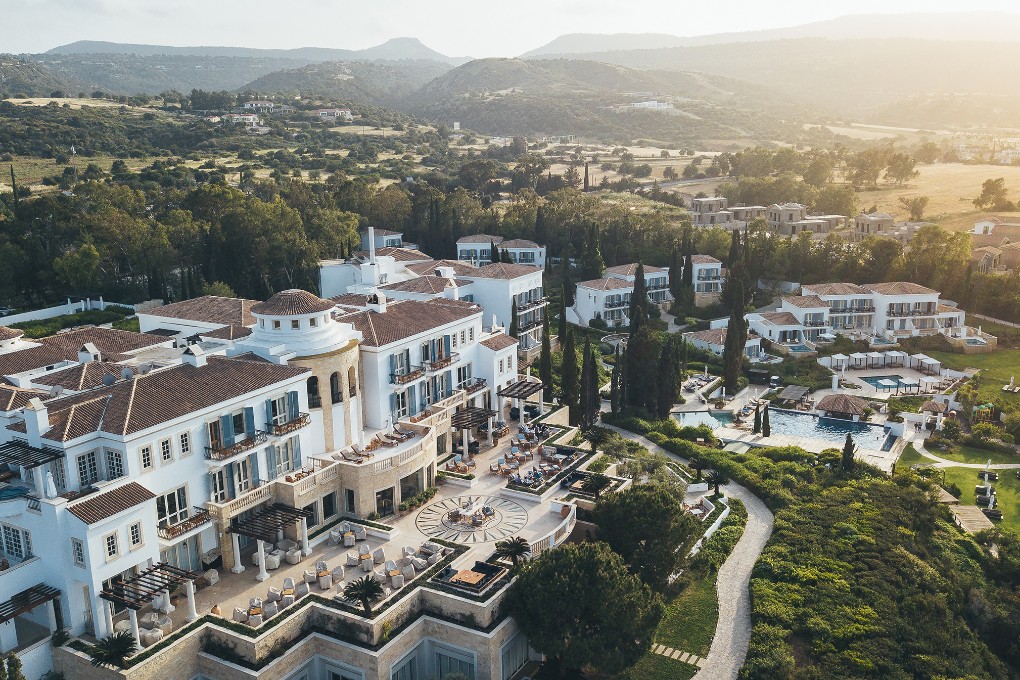 Aerial view of Anassa hotel in Cyprus featuring a white walled and tiled roof building surrounded by lush gardens with small villages and hills behind