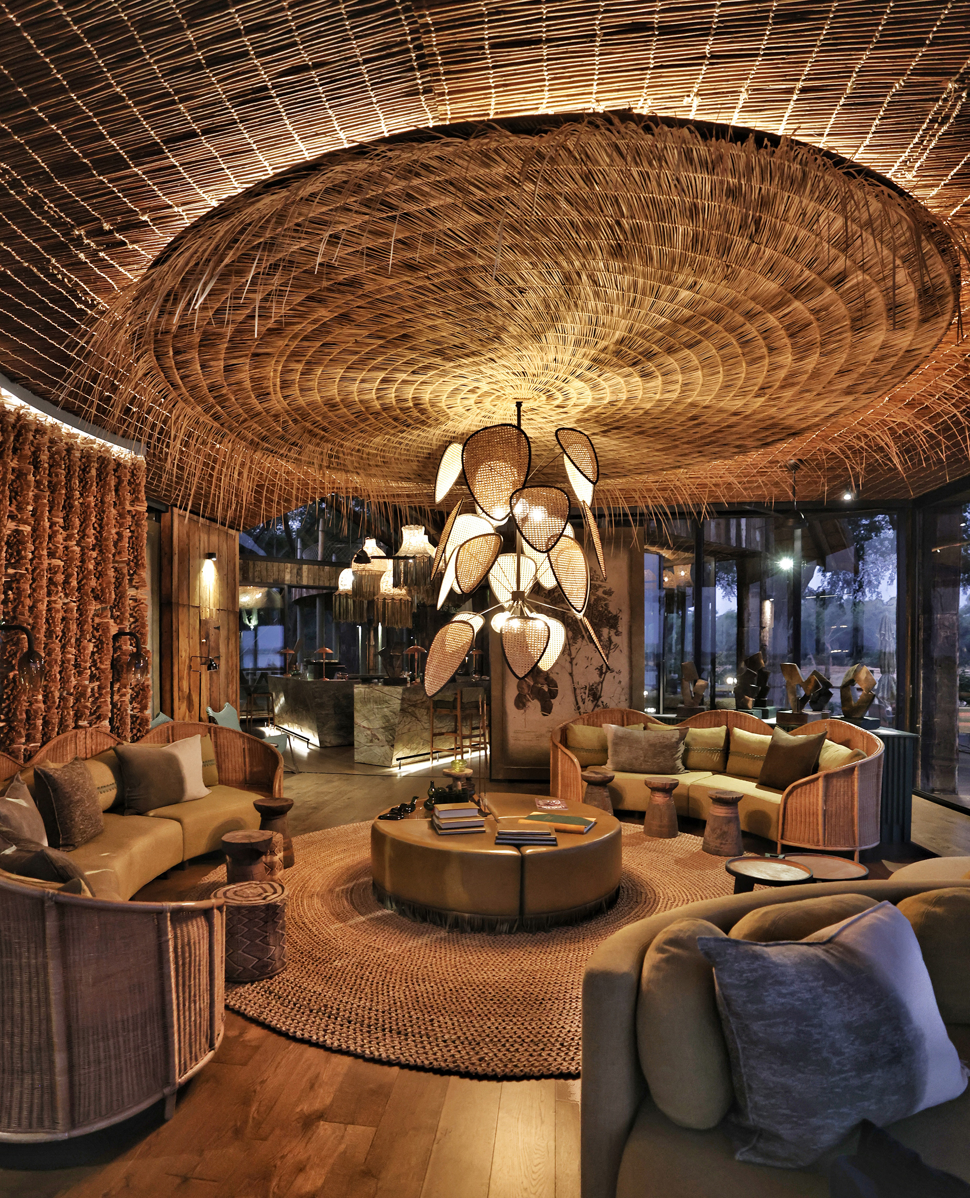 A cosy lounge lit at dusk with a circular coffee table surrounded by sofas below a statement light fixture