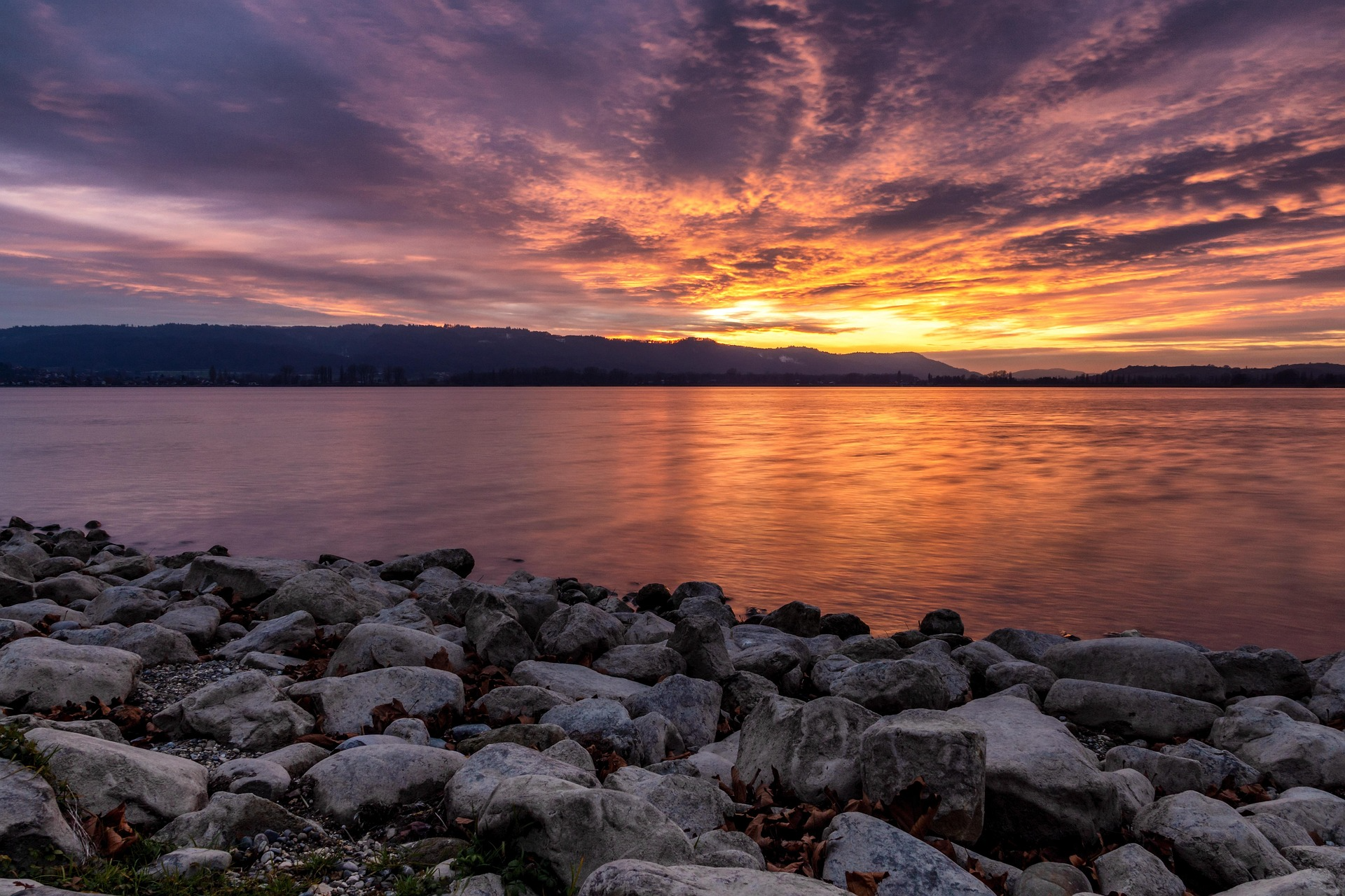 Rocky lakeshore in the foreground with calm water reflecting a vivid orange and purple sunset over distant hills.