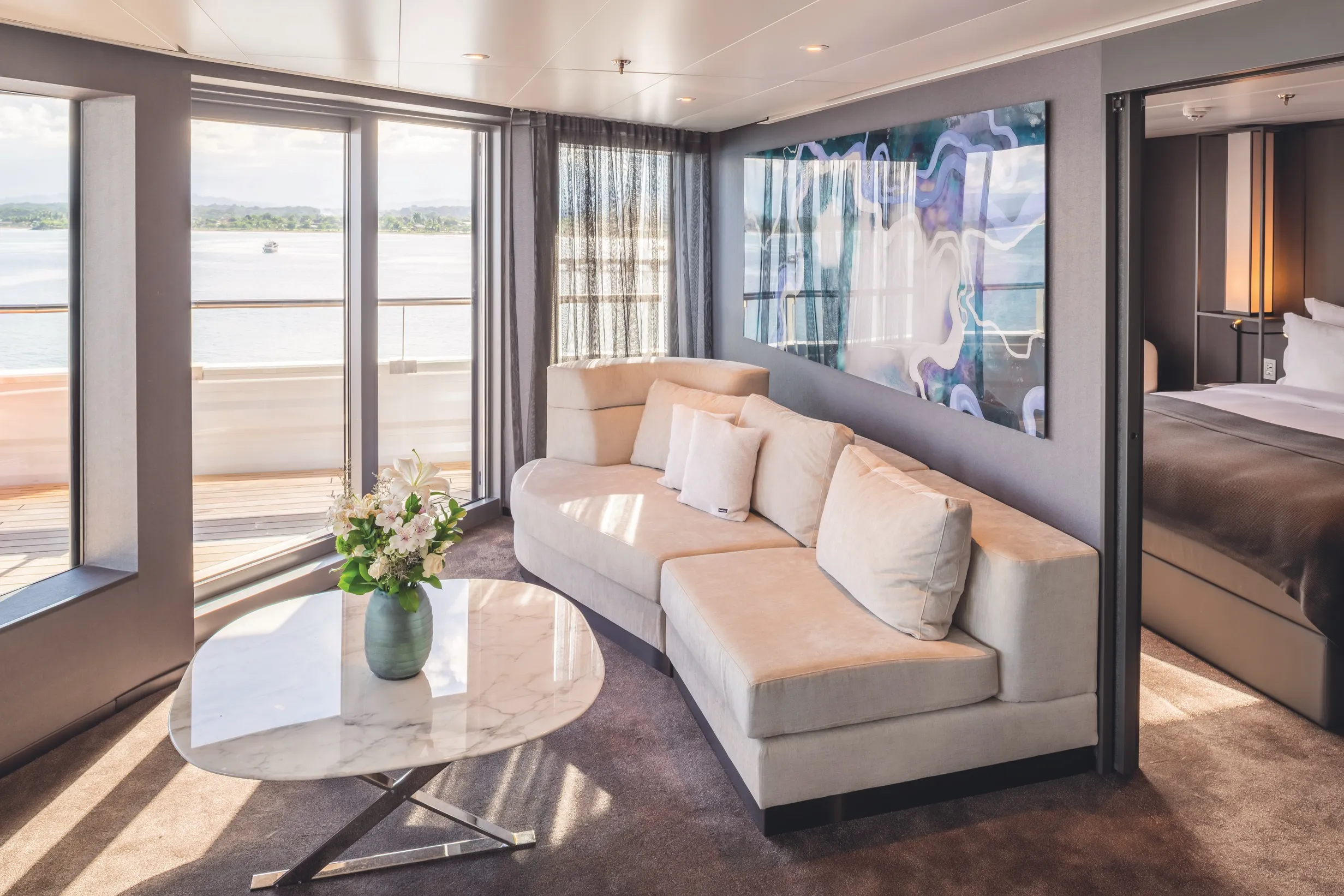 Modern living room with beige sectional sofa, marble coffee table, and large windows overlooking the water.