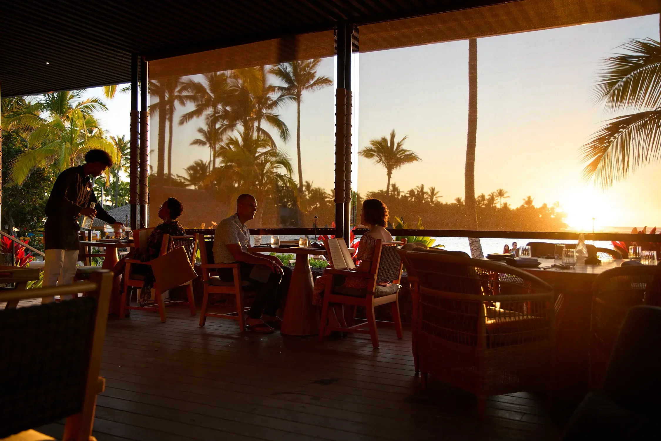 Guests eating in Moana Restaurant at Rosewood Kona Village slightly silhouetted against the blinds with a sunset behind