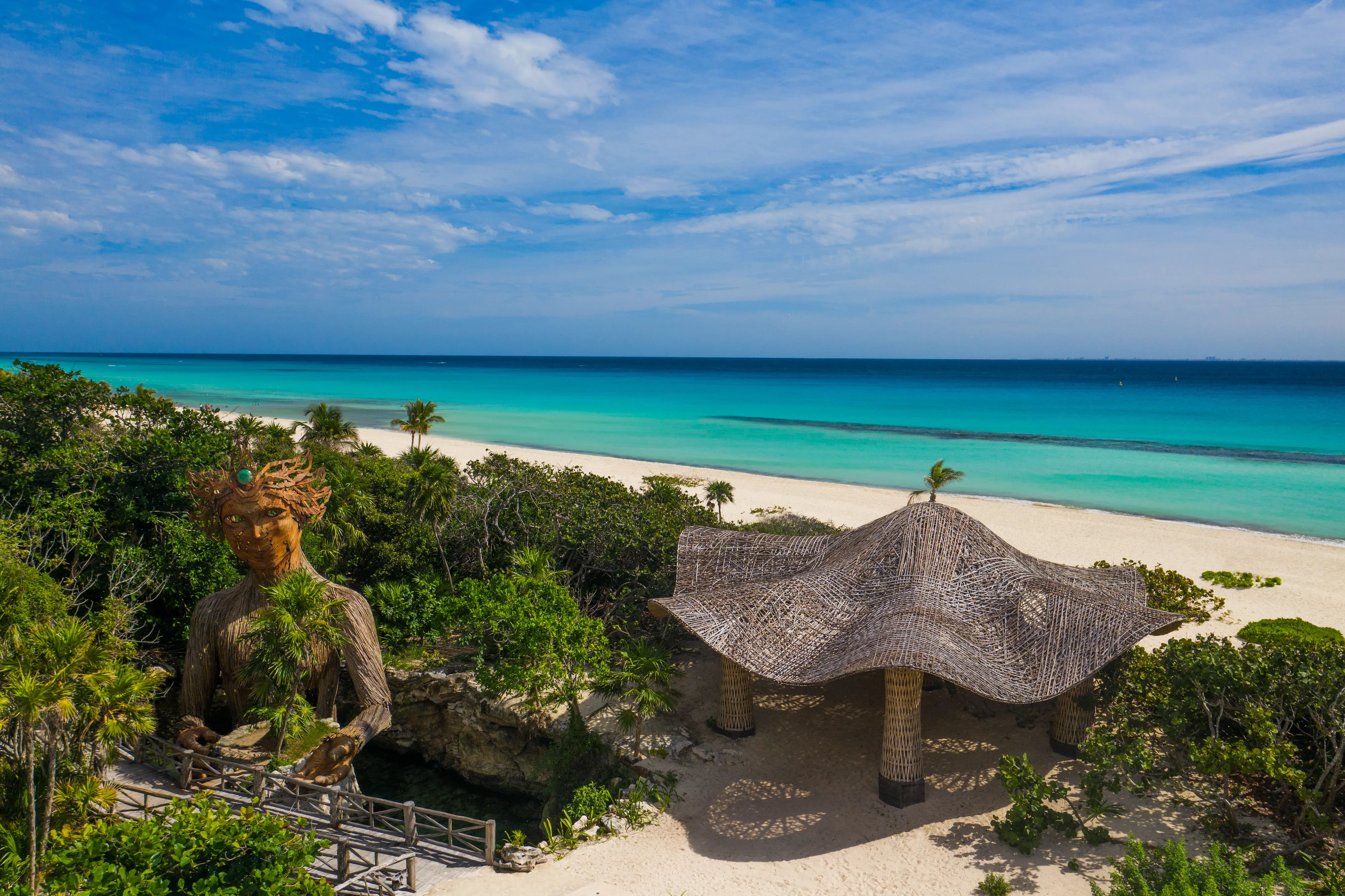 A thatched mushroom-shaped pavilion and tall female statue surrounded by lush greenery on a pristine beach beneath a clear sky with the ocean in the background.