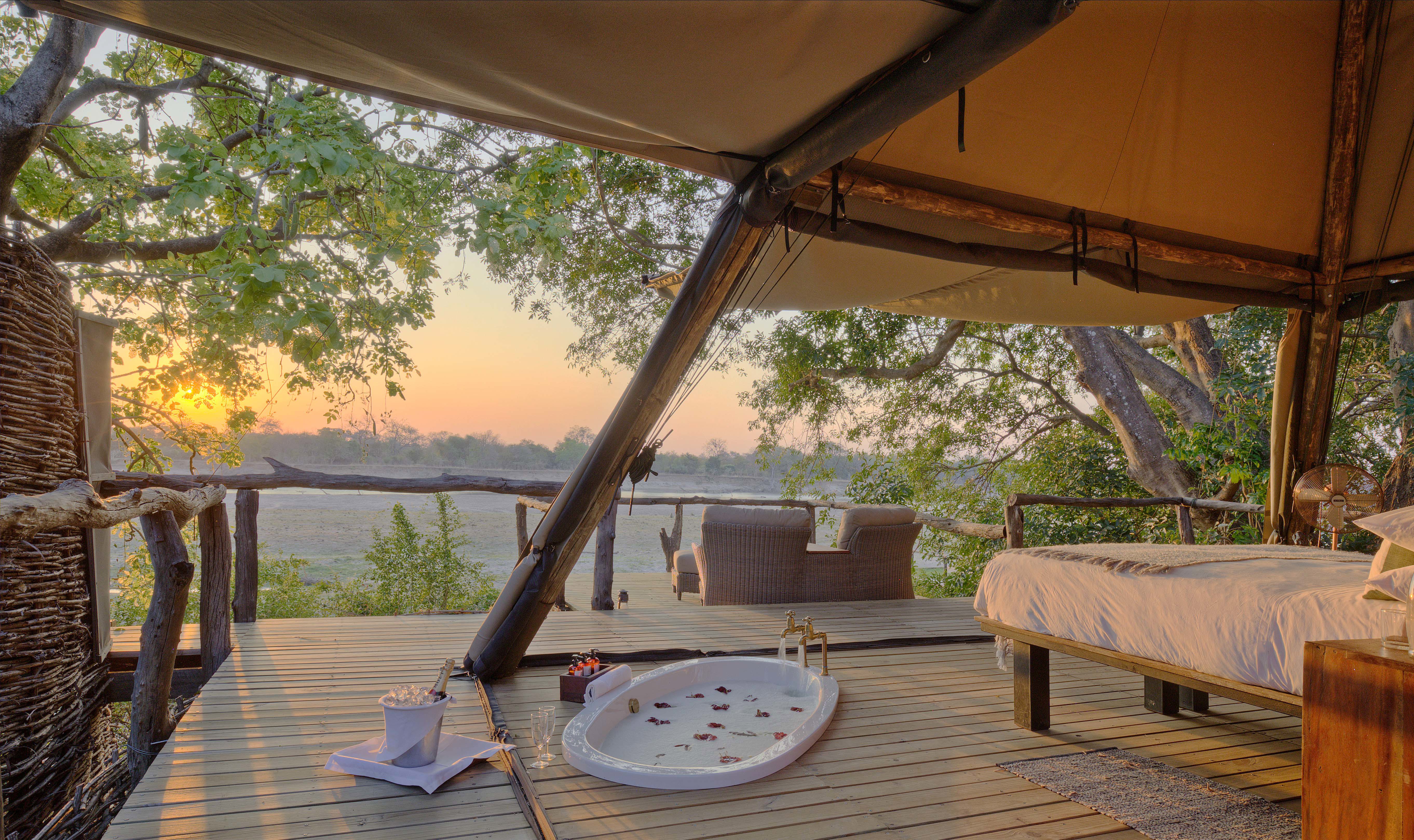 A luxurious open-air tented room at Shawa Luangwa Camp in Zambia, featuring a bed, a bathtub with rose petals, and an outdoor seating area overlooking a scenic view of trees and the sunset.