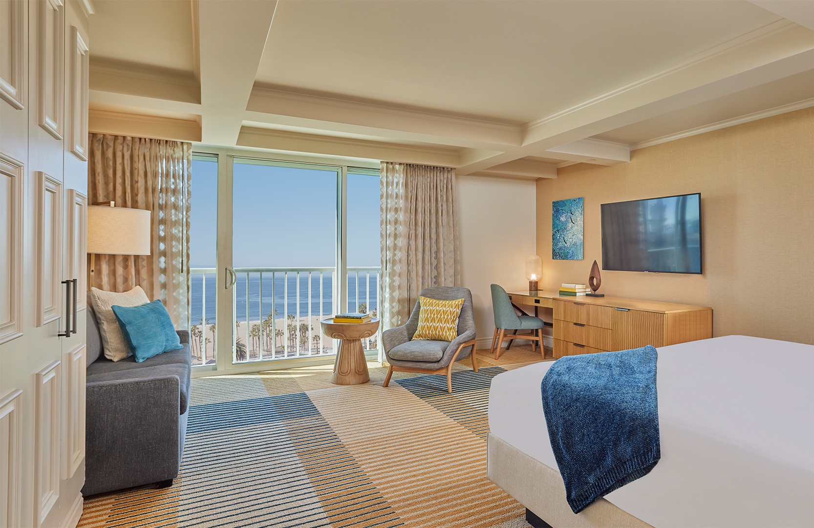 A hotel suite with a window view of a blue sky and ocean. The room is decorated in cream and blue tones and features a plush double bed, a large chest of drawers and desk area, a TV and plush seating areas.
