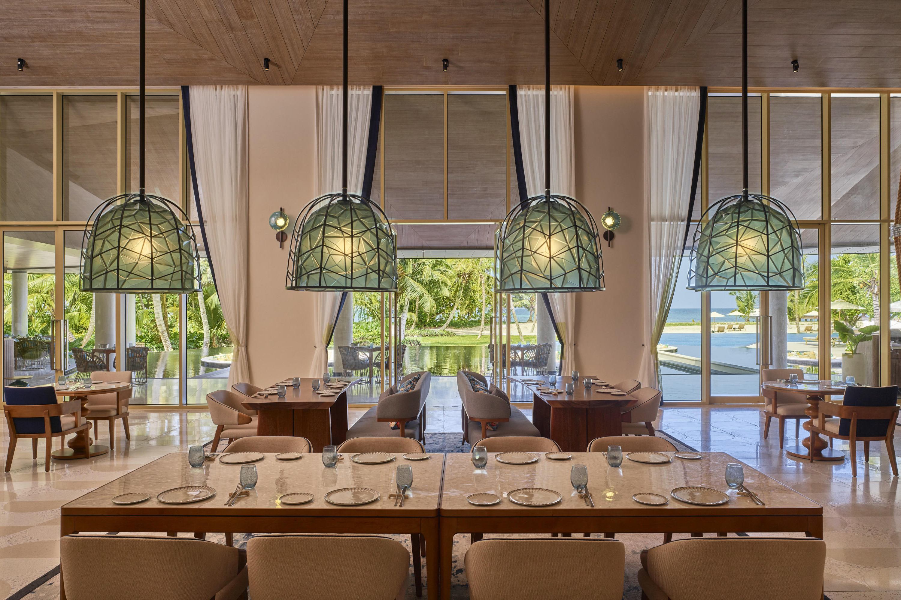 A large table in Saba La Perle restaurant set with china and framed by geometric green light shades with a view of the pool out of the double doors in the background