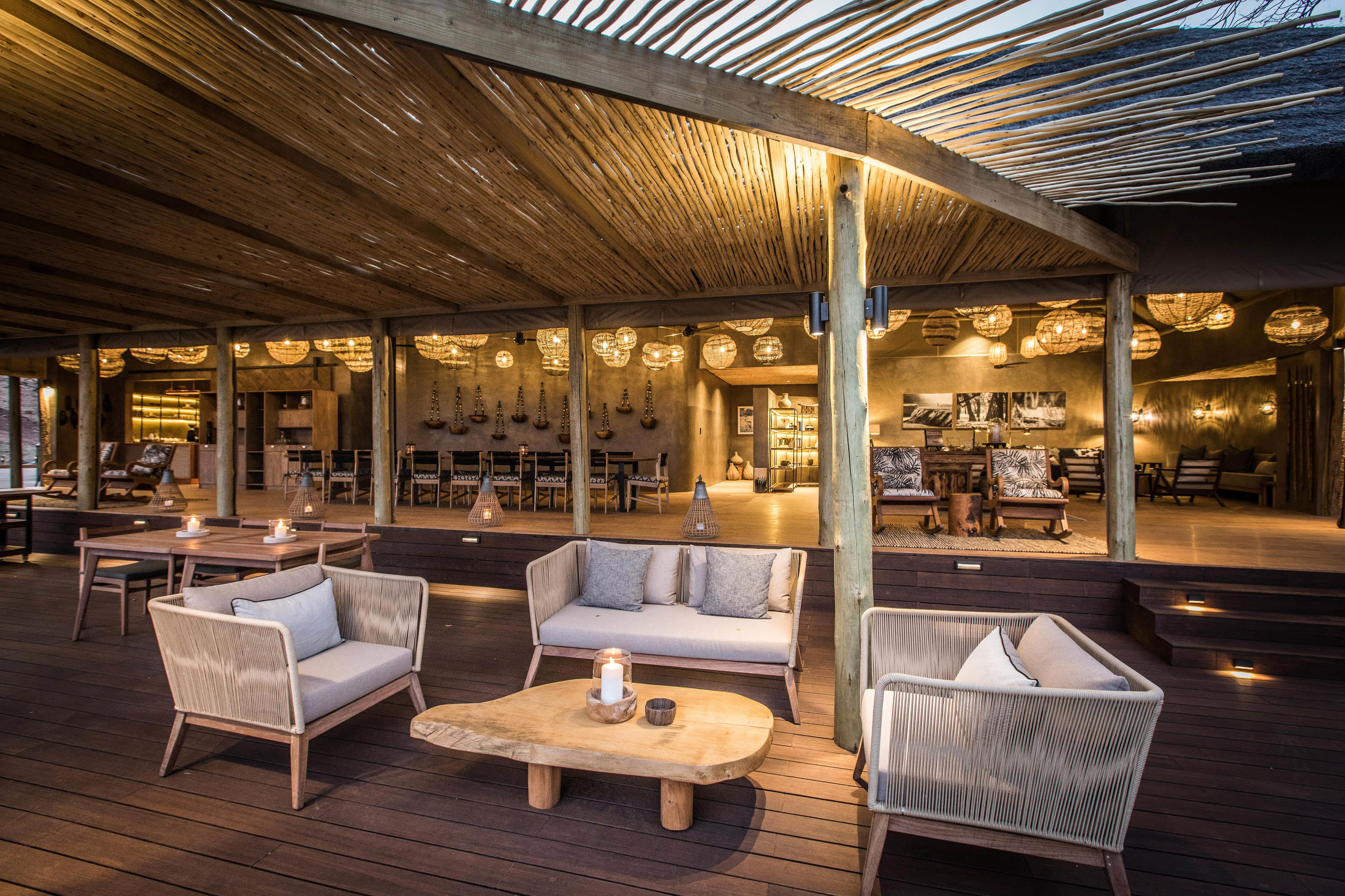 Iutdoor lounge area at Puku Ridge Camp with wooden furniture, cushioned seating, and a rustic roof, with an open-air dining space featuring hanging lanterns and warm lighting in the background