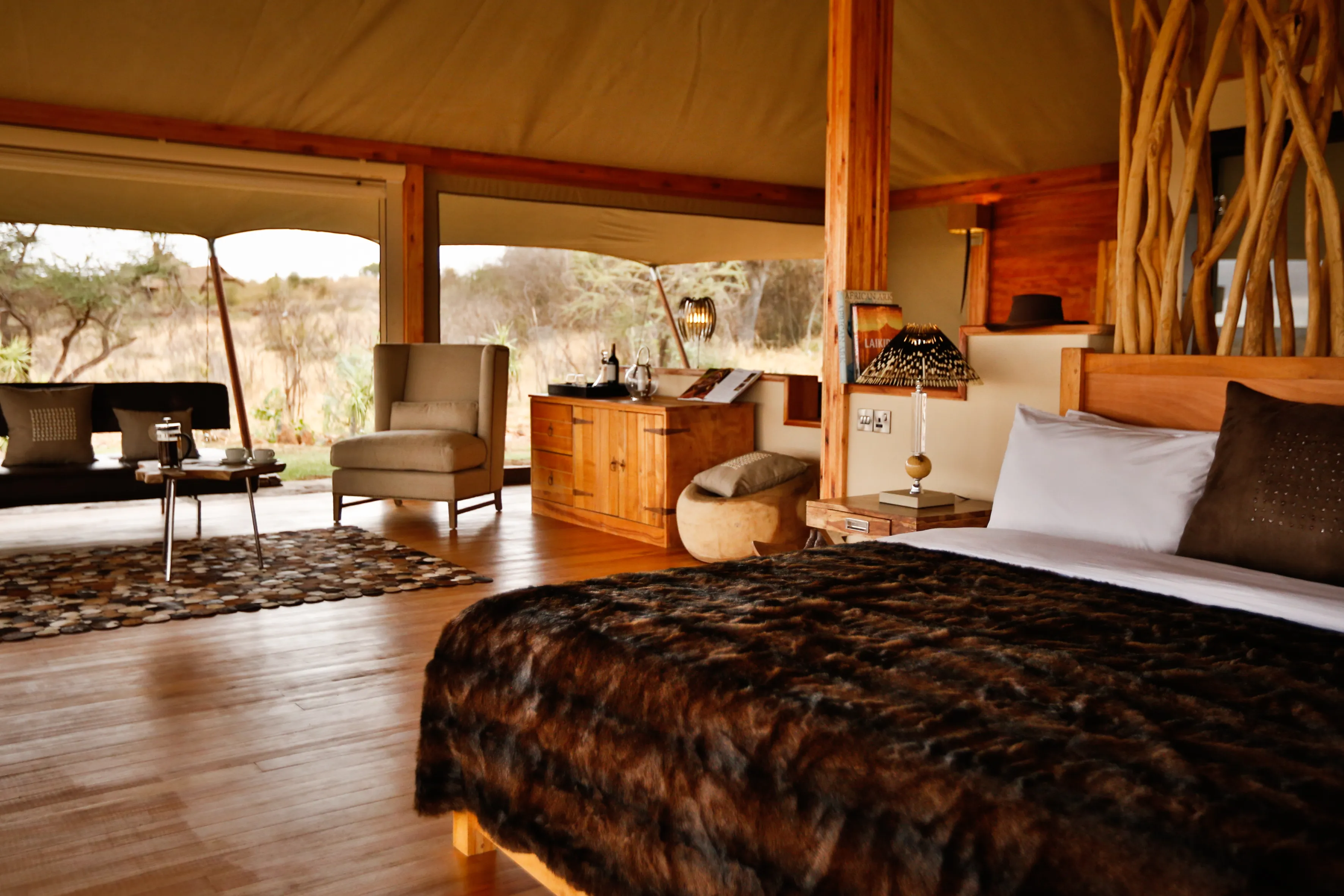 The interior of a double tent suite at Elewana Loisaba Tented Camp featuring a comfy bed with a dark throw, a small lounge area and cabinet with a view of a grassy landscape behind
