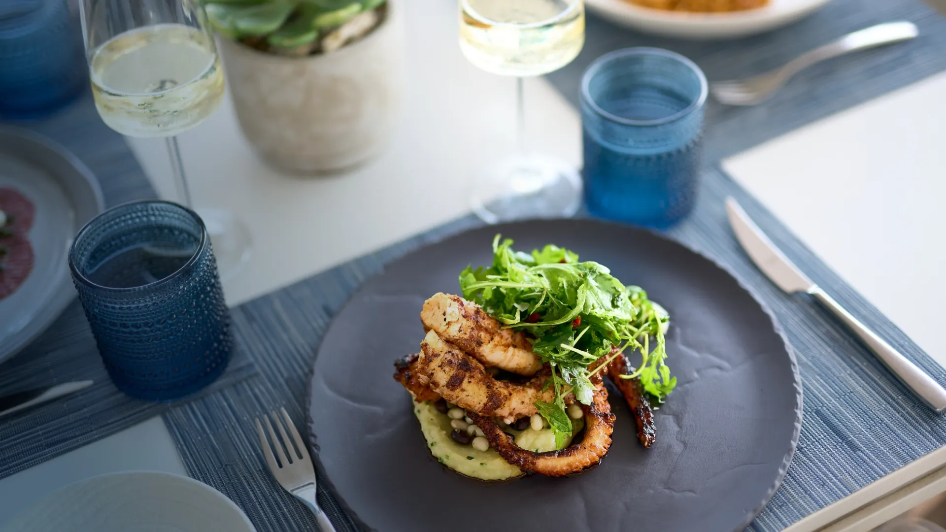 A gourmet dish of grilled octopus and greens on a black plate, with blue glassware and wine glasses on the table.
