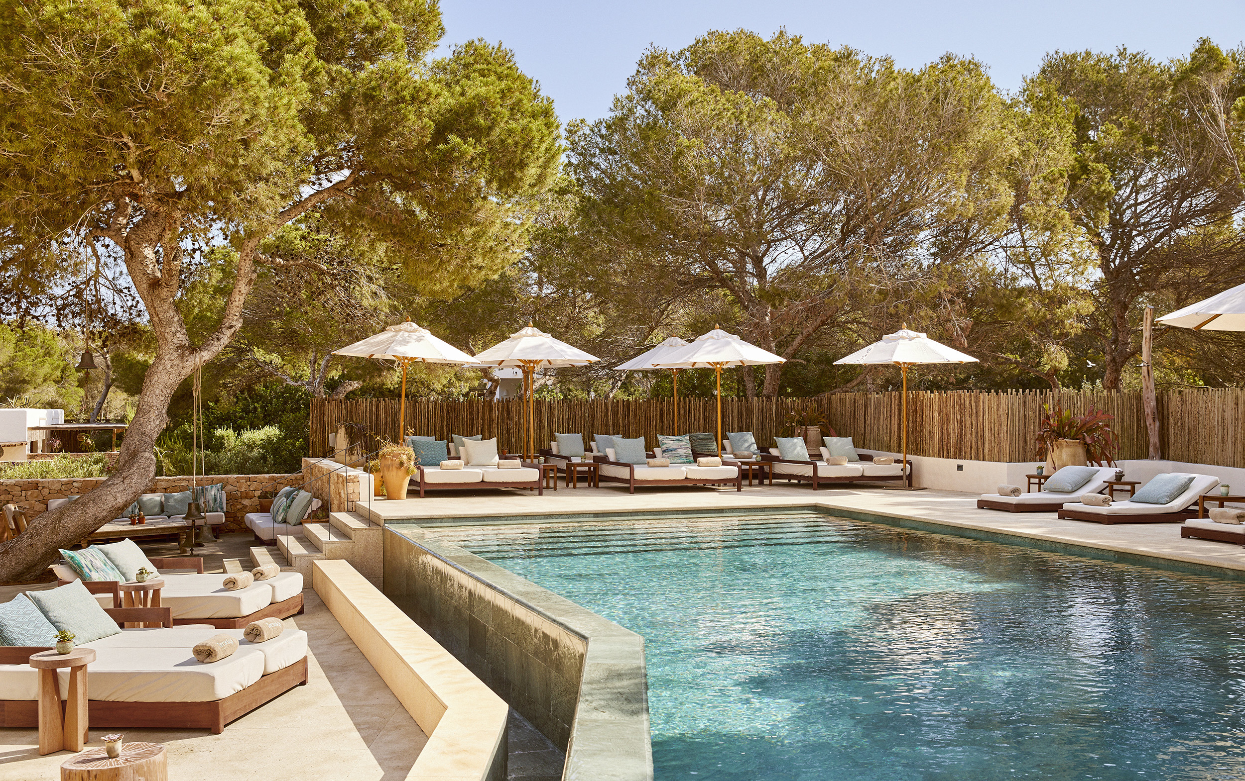 A relaxed pool area with sun loungers decorated with blue cushions set around a pool beneath Mediterranean trees
