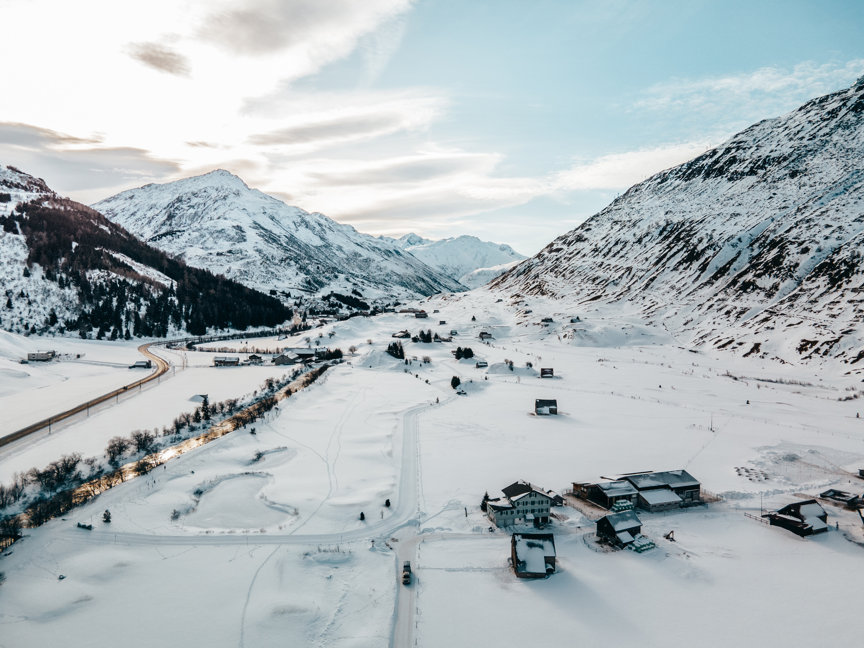 Landscape of Andermatt in Switzerland with snow-covered valley, mountains, buildings, and a winding road.