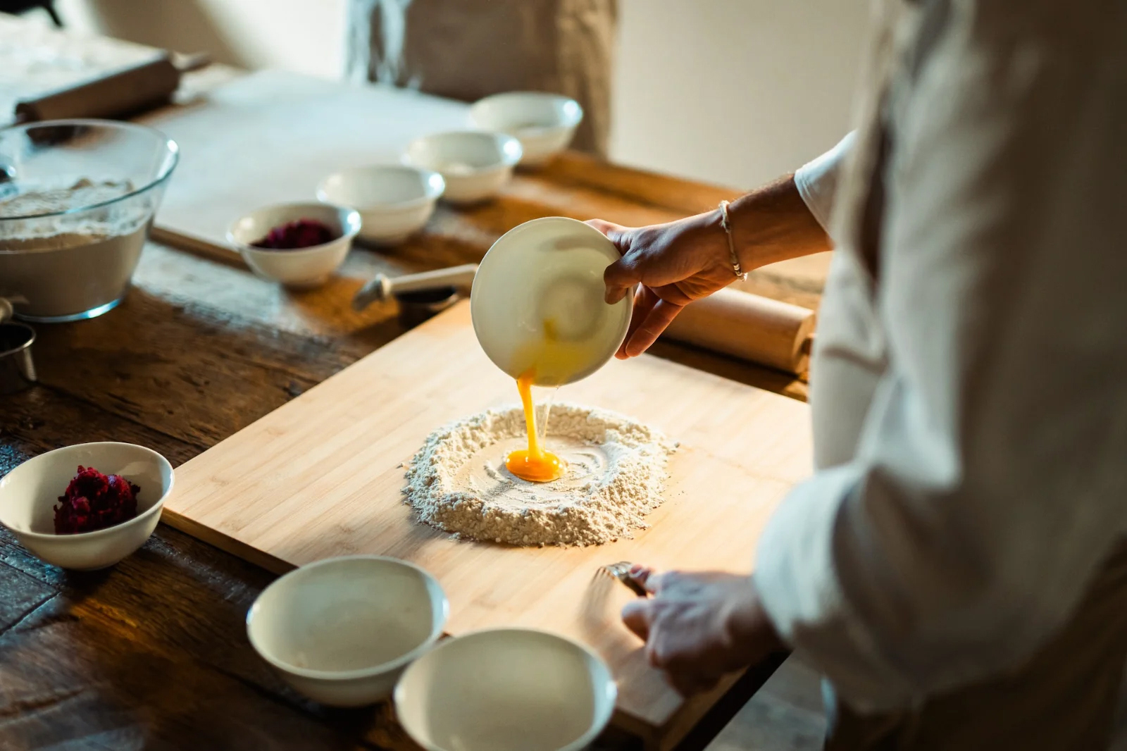 Chef in white pouring scrambled egg into flour to make pasta on a large wooden board surrounded by bowls