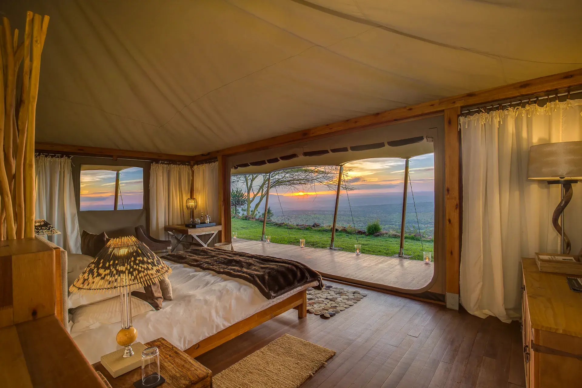 A luxury tent at Elewana Loisaba Tented Camp featuring a comfy bed and African-inspired furniture beneath a big canvas roof with a large opening looking out over the landscape at sunset