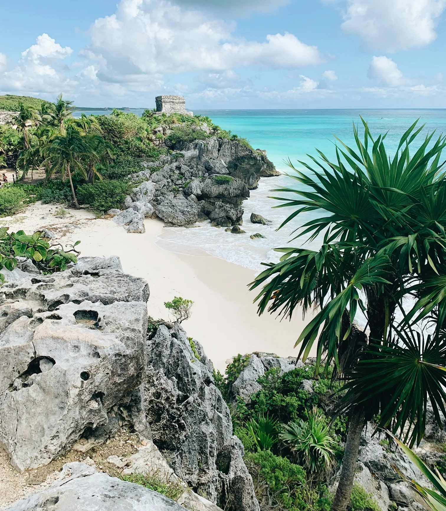 A tropical beach with turquoise waters, rocky cliffs, lush greenery, and a small ancient stone structure on a cliff.
