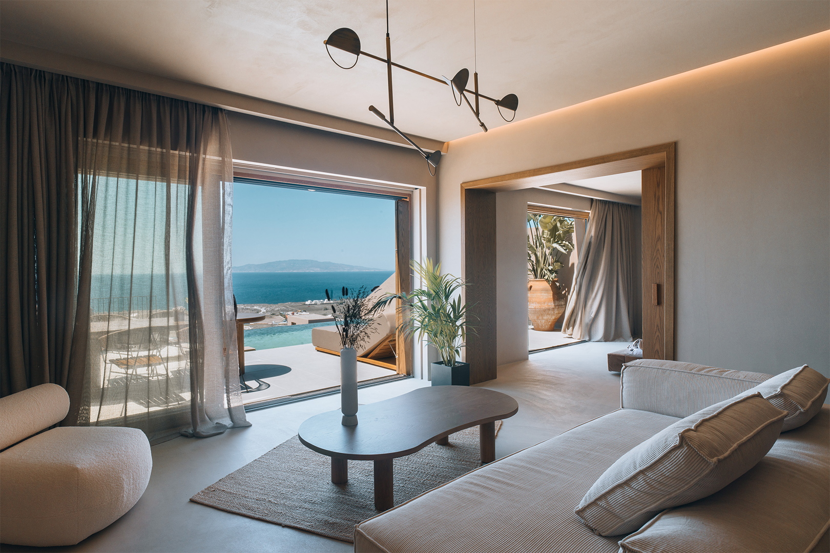 An airy hotel living room with sofa, plush chair and coffee table is decorated in light collours and wood tones, its patio doors looking out over the sea