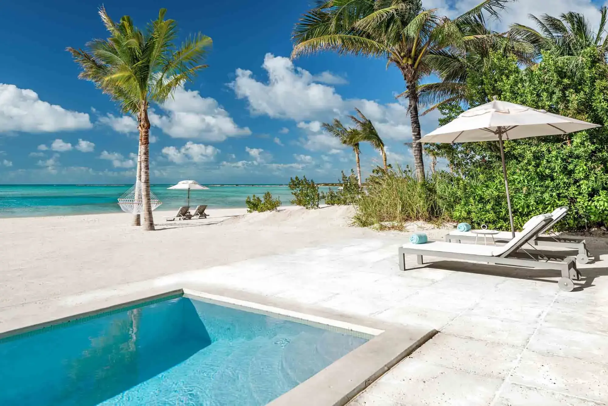 Sleek pool terrace of Beachfront Pool Suites Pool at Ambergris Cay in Turks & Caicos featuring a plunge pool and loungers on a pale tiled terrace at the side of a white sand beach with palms
