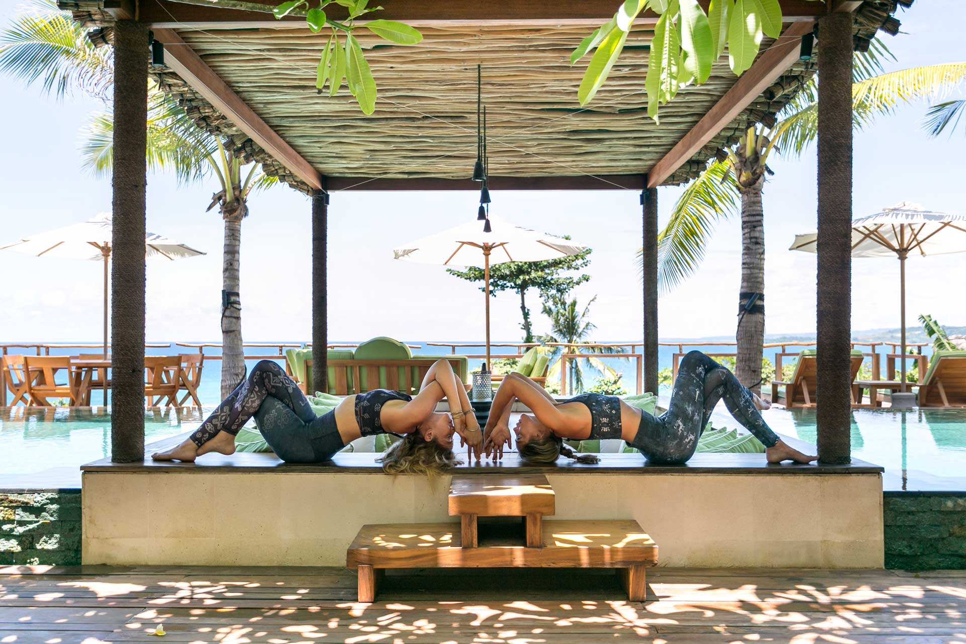 Two people practising mirrored yoga poses on a platform beside a pool with palm trees and sea views in the background.