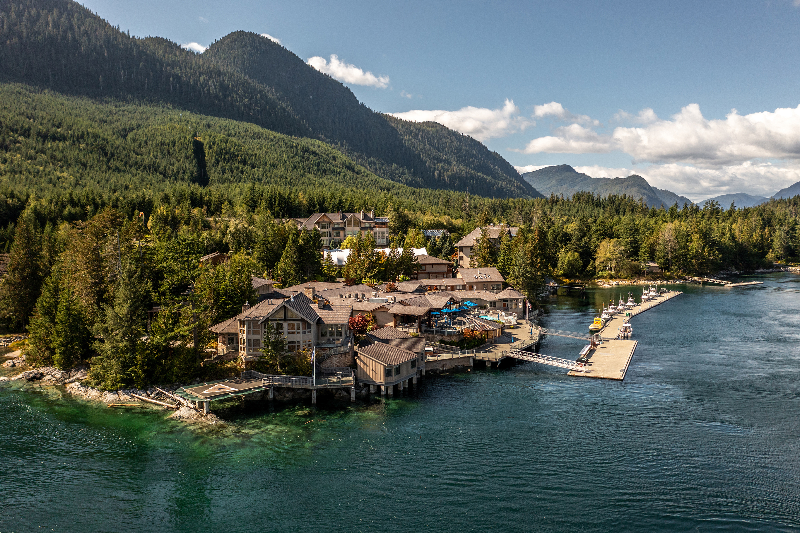 A large wooden resort by a lake surrounded by trees with mountains in the background