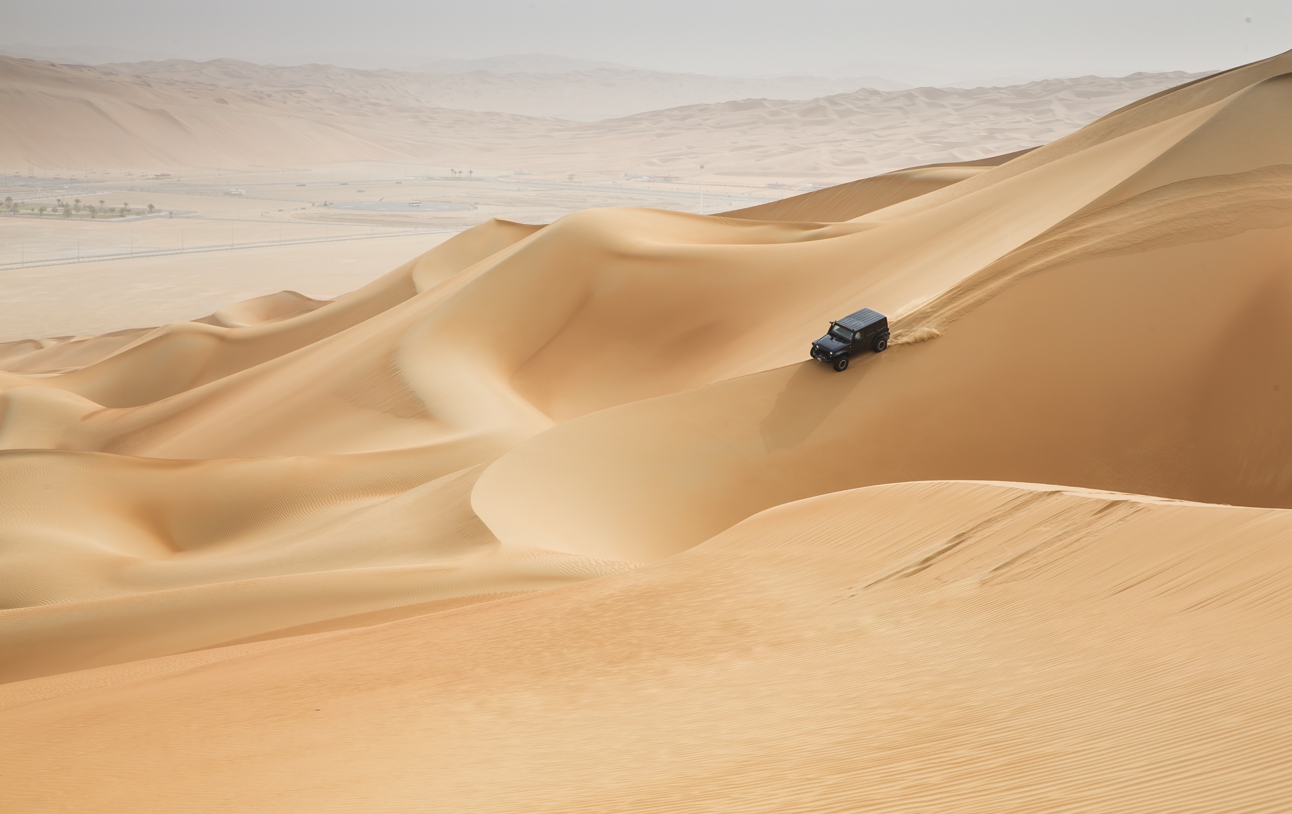 A black SUV drives down a large sand dune in a vast Arabian desert landscape.