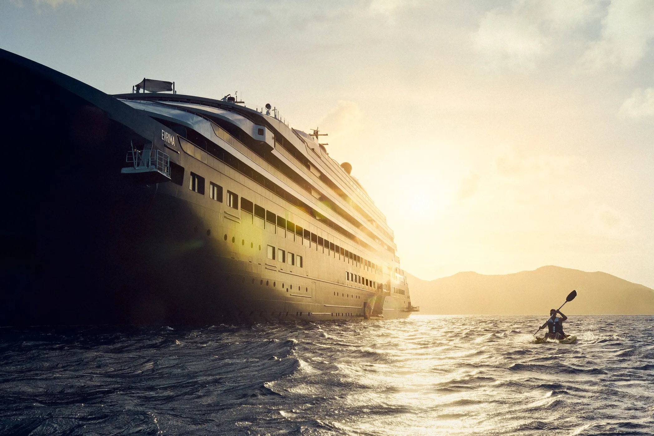 A large cruise ship sailing on the ocean at sunset with a person kayaking nearby.