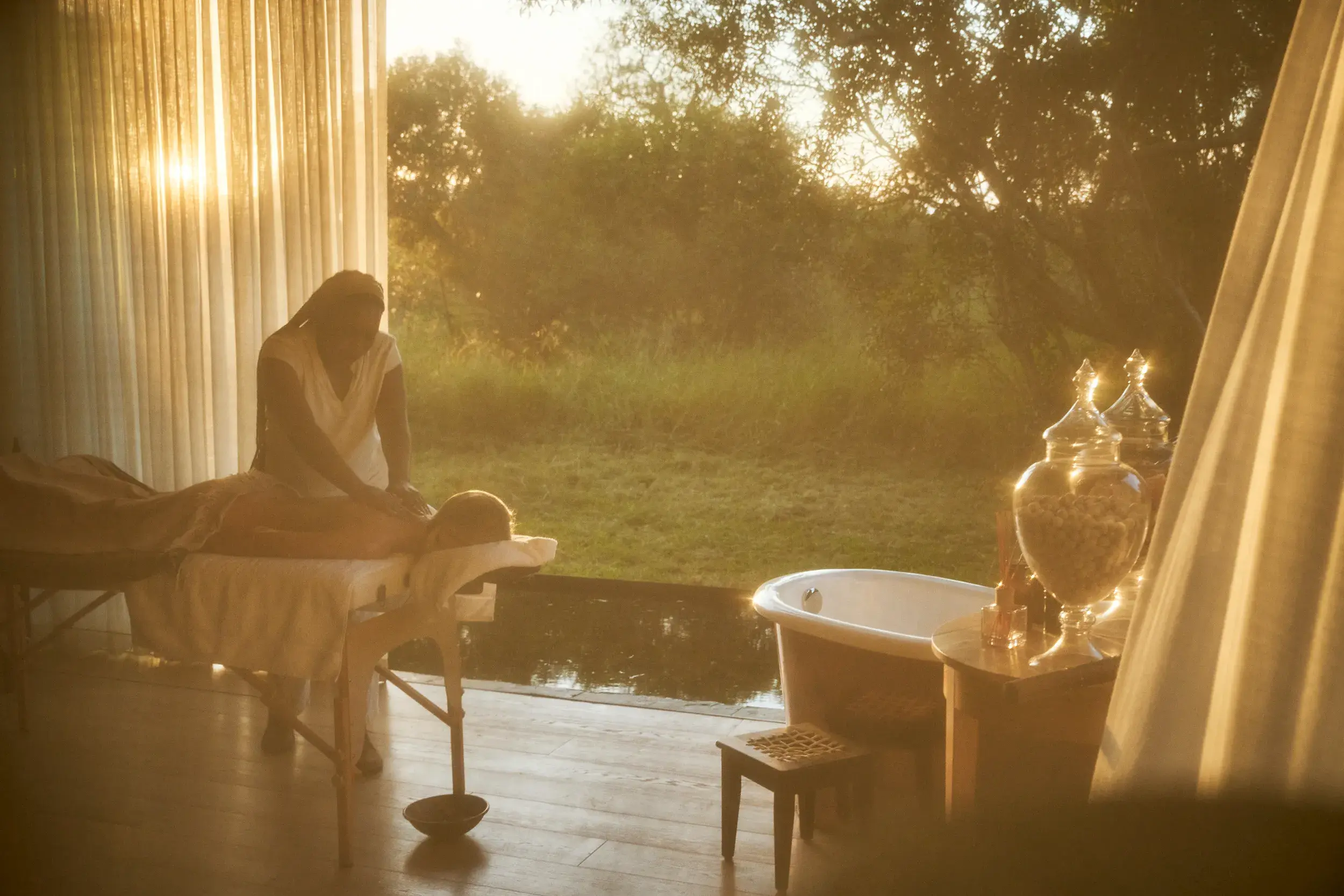 A person receiving a massage in a peaceful building with a large open wall looking out onto a pool and trees, with jars of items and a deep bath to the side