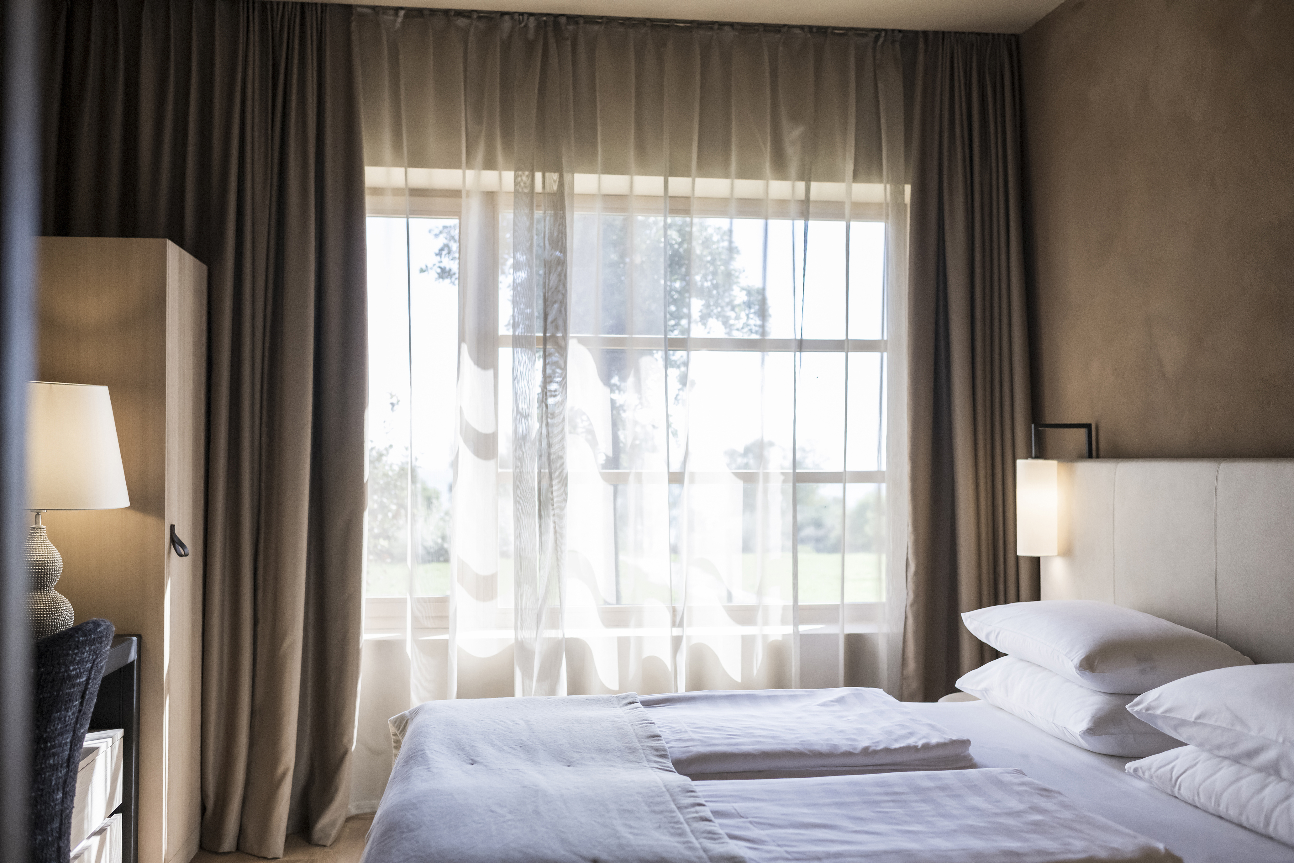 An airy bedroom with white linens, pale wooden furniture and sheer white curtains drawn across big windows 