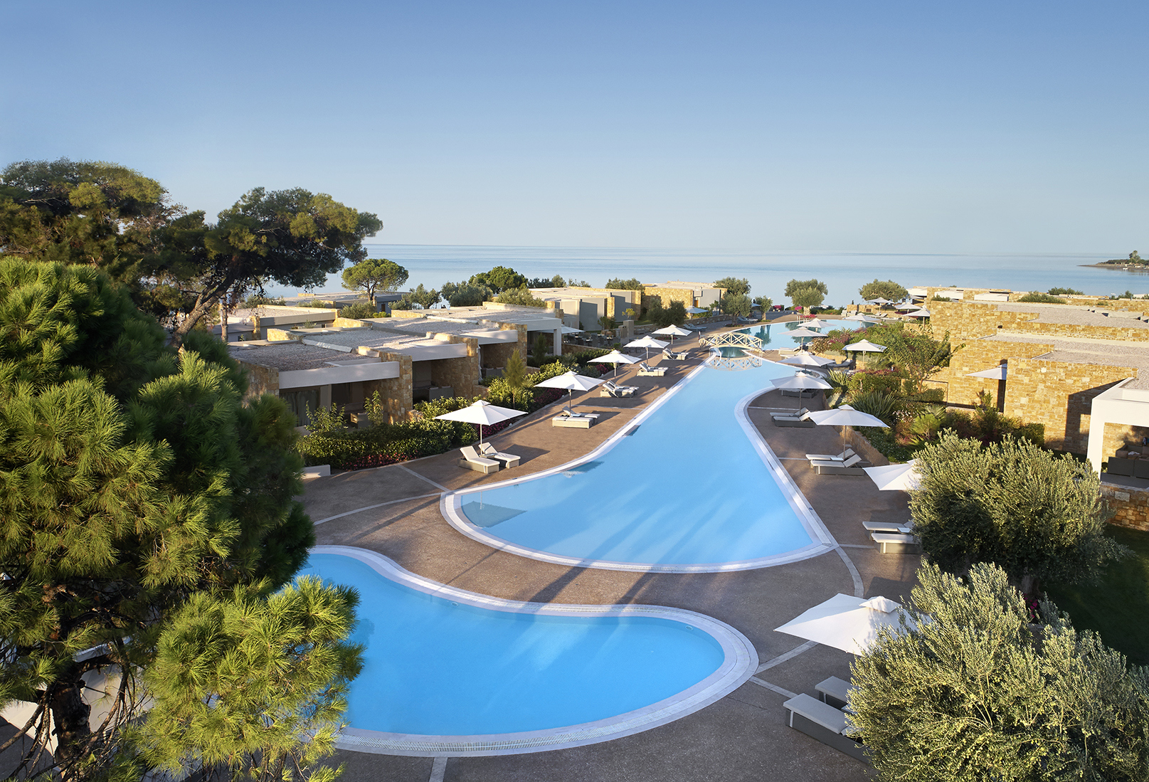 Two asymetrical swimming pools in a space outside stone suites surrounded by trees with the sea in the background