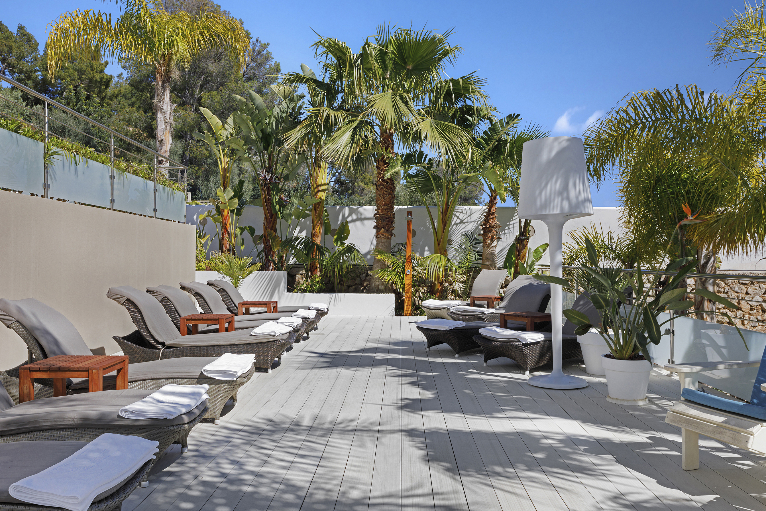 Luxurious outdoor lounge area at Portals Hills in Mallorca with wicker sun loungers, palm trees, and a large white lamp under a clear blue sky.