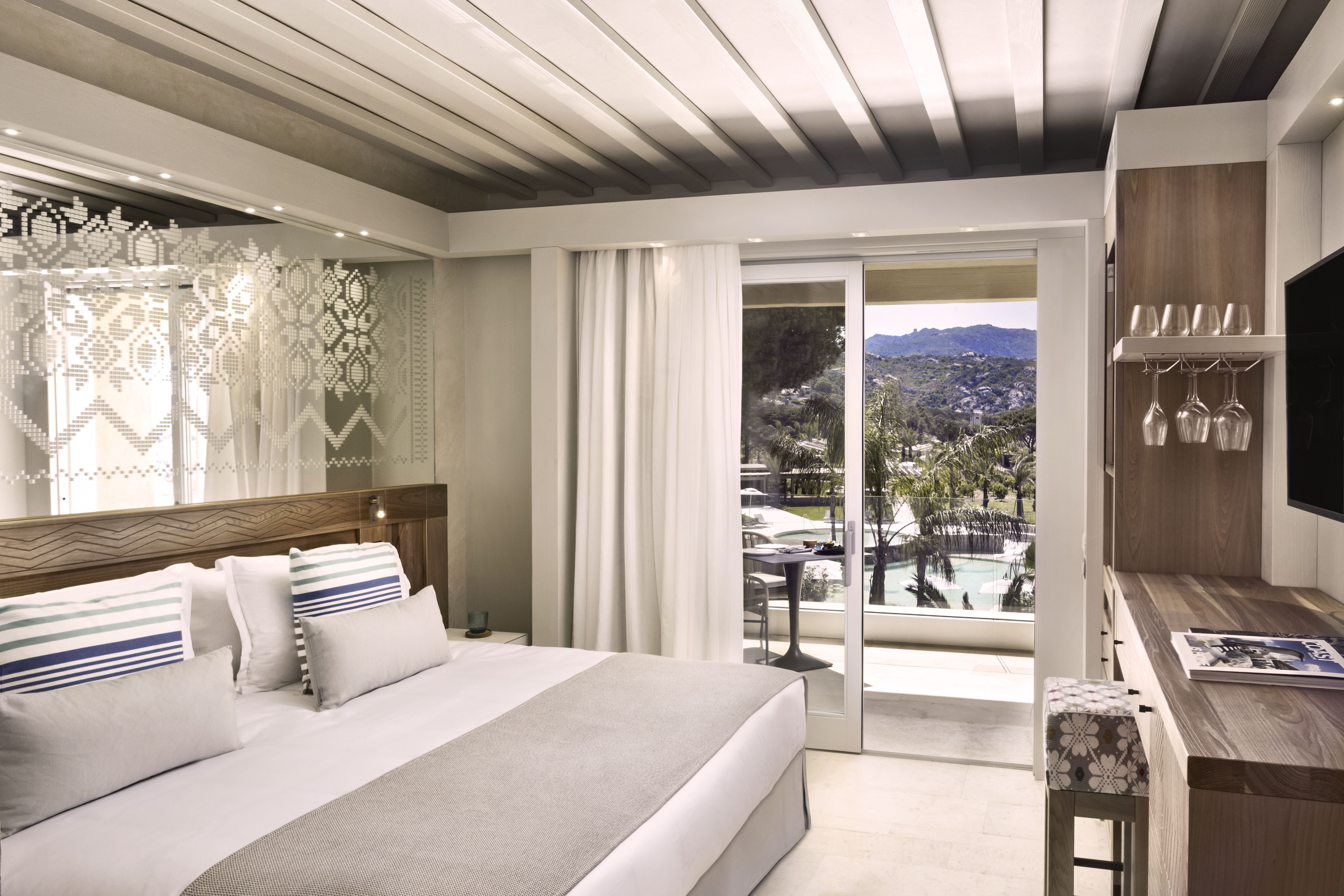 A sleek suite featuring a bed and desk area with sliding door onto a balcony overlooking a pool