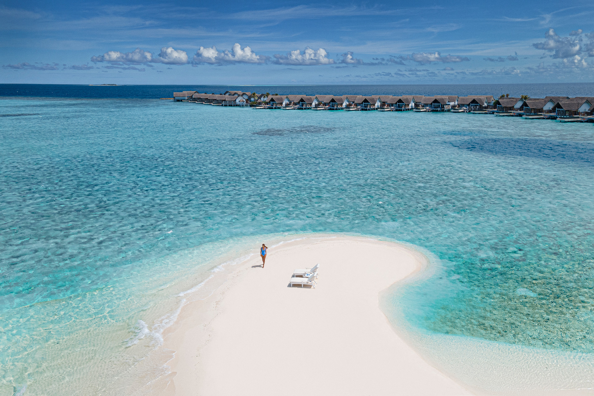 A woman stoof on a sand bank with two sun loungers and overwater villas in the background