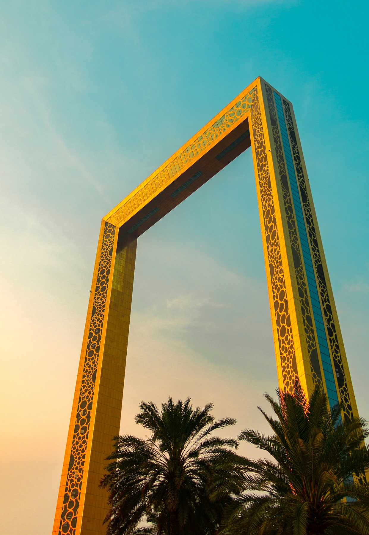 A tall, golden rectangular structure with intricate circular patterns, known as the Dubai Frame, stands against a clear blue sky, offering panoramic views of the city for families exploring modern landmarks.