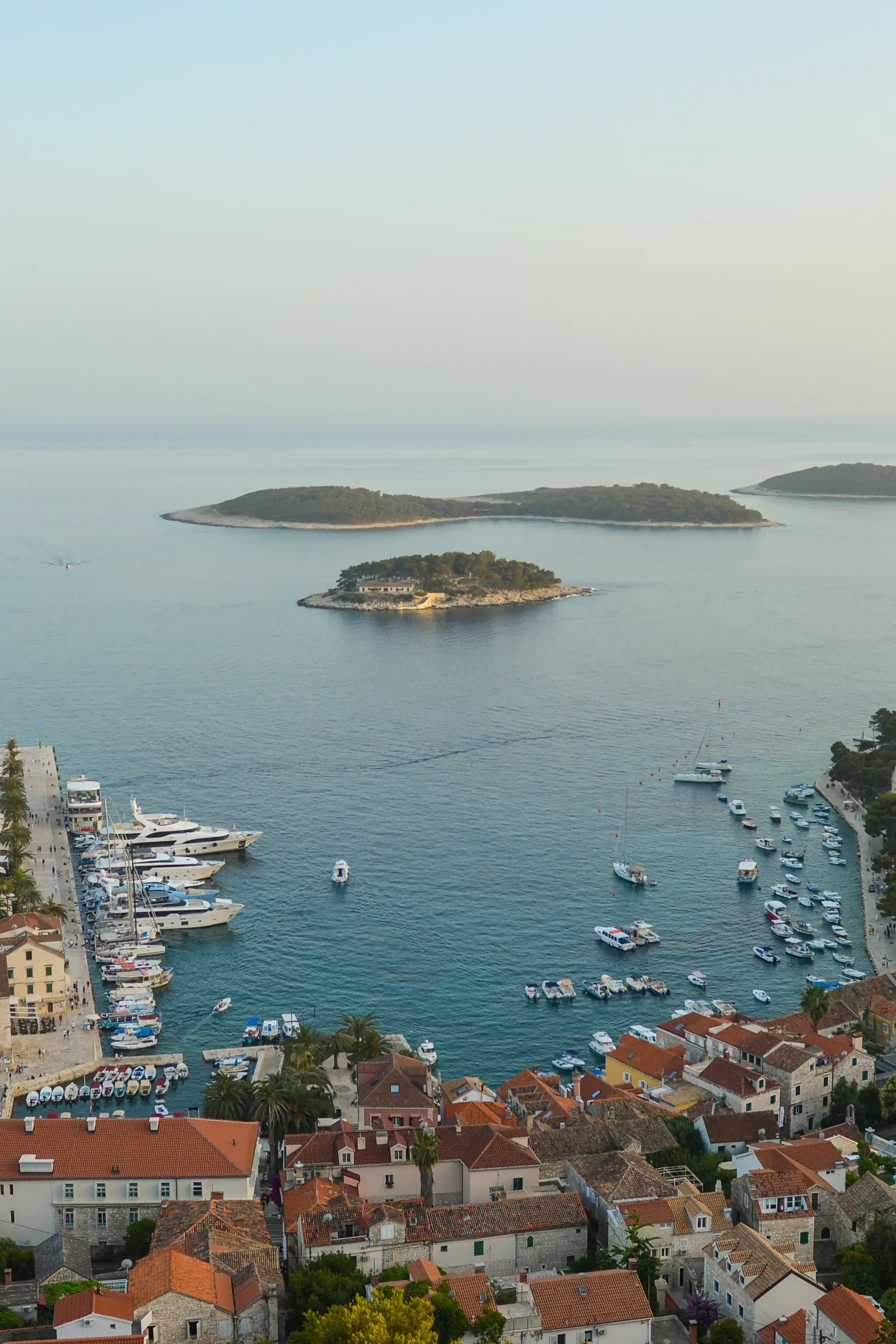 An elevated view of Hvar harbour dotted with yachts, with the Pakleni Islands further out to see under a hazy sky