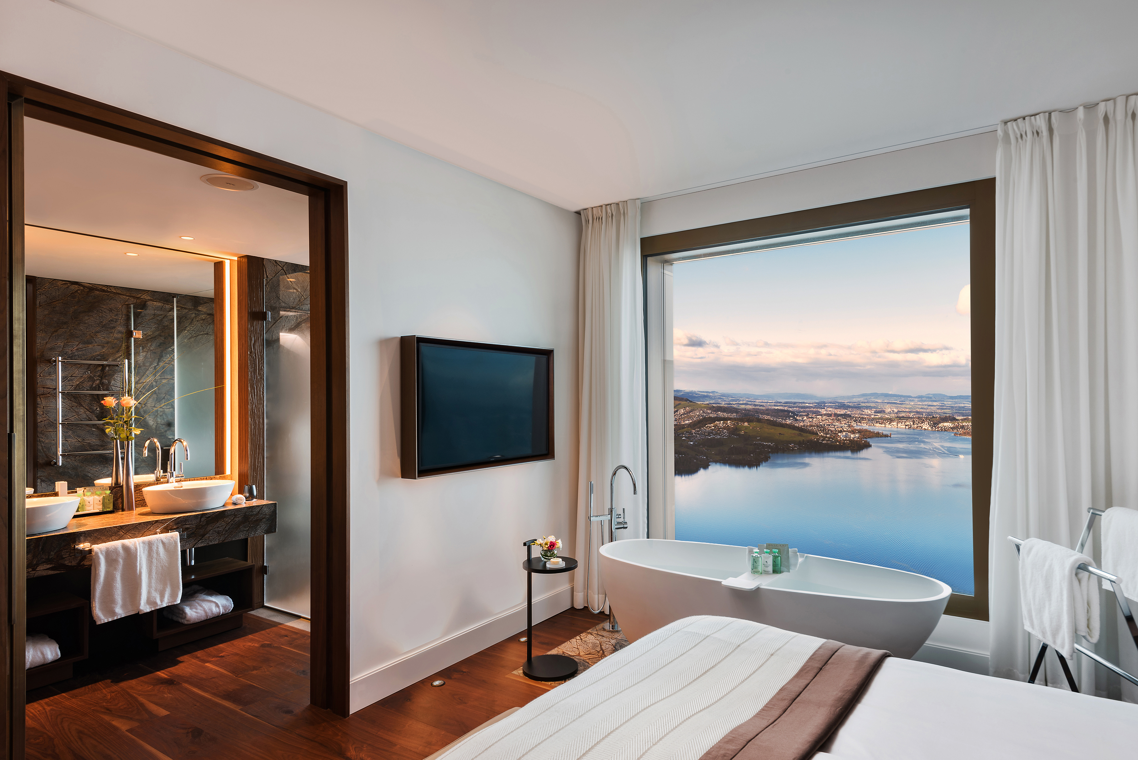 An airy Premium Suite at Bürgenstock Hotel showing the corner of a bed and a deep bathtub in a large square window with a lake view, with a glimpse of the bathroom beyond
