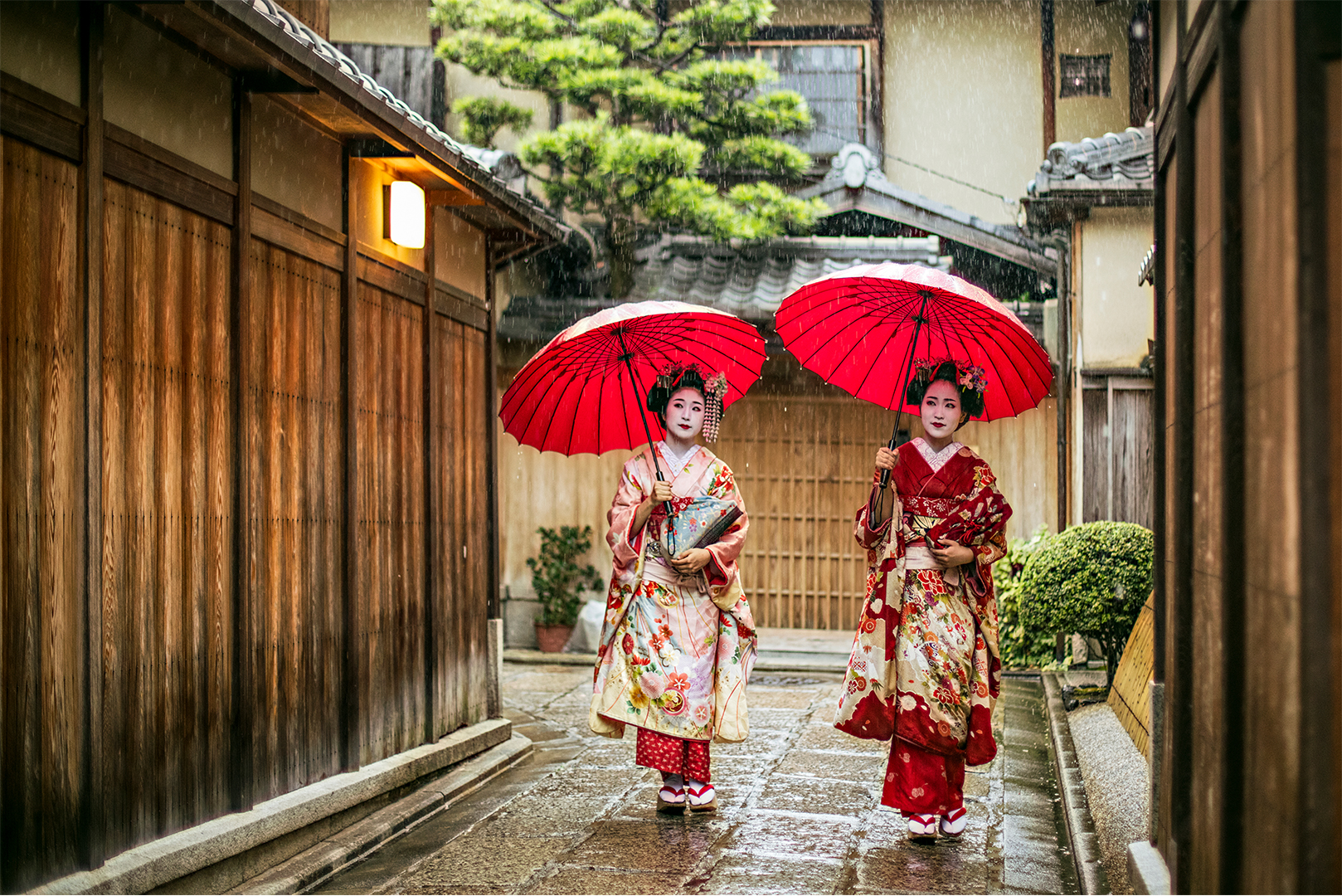 Two individuals in traditional Japanese kimonos holding red umbrellas on a rainy day in a historic alley.