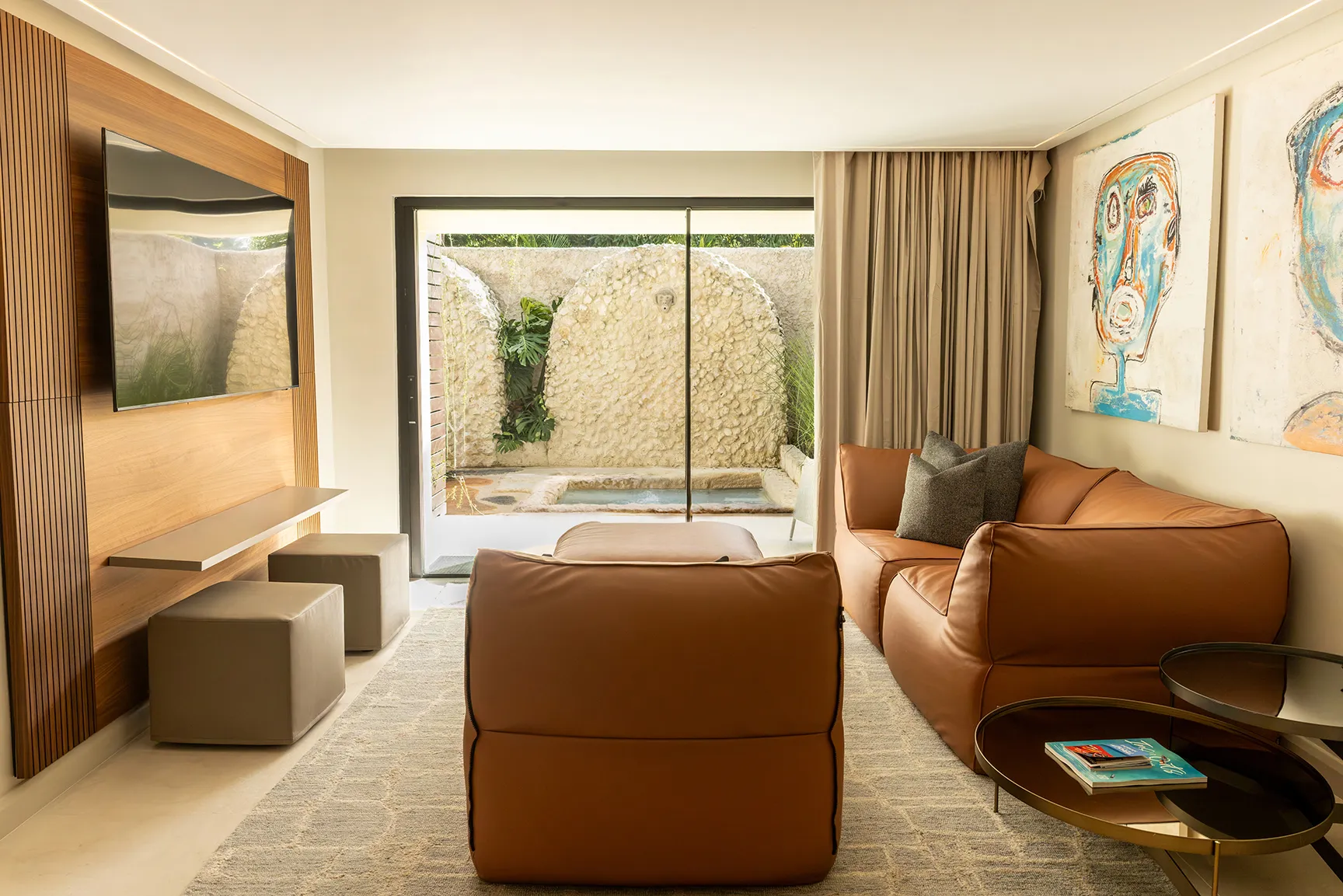 Modern suite living area with leather seating, large TV, abstract artwork, and sliding doors opening to a private patio with a plunge pool.