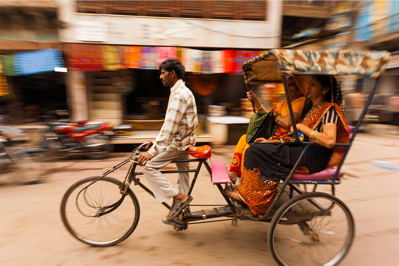 A person pedalling a rickshaw with a passenger on board, in front of blurred colourful storefronts, suggesting motion.
