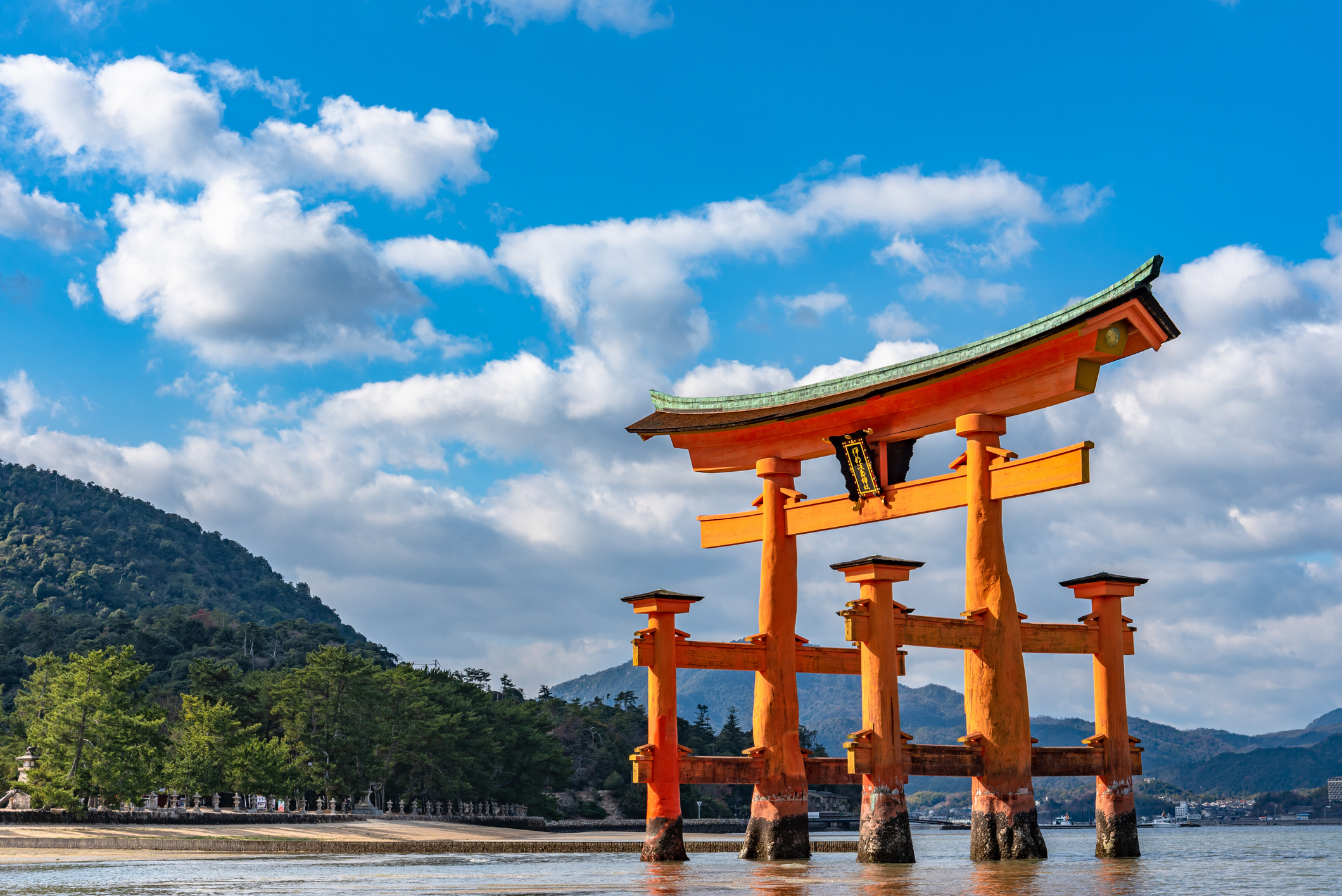 A large, traditional Japanese torii gate stands in shallow water with a mountain and blue sky with clouds in the background.