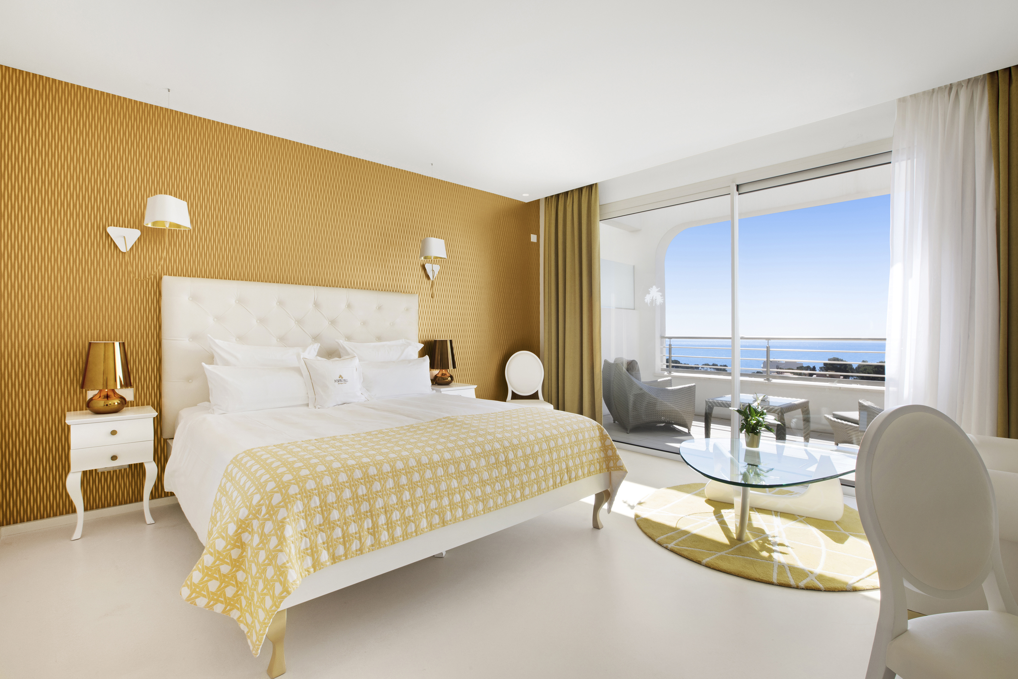 Luxurious hotel room at Portals Hills in Mallorca with elegant white furniture, a yellow-patterned bed, and a balcony overlooking the ocean.