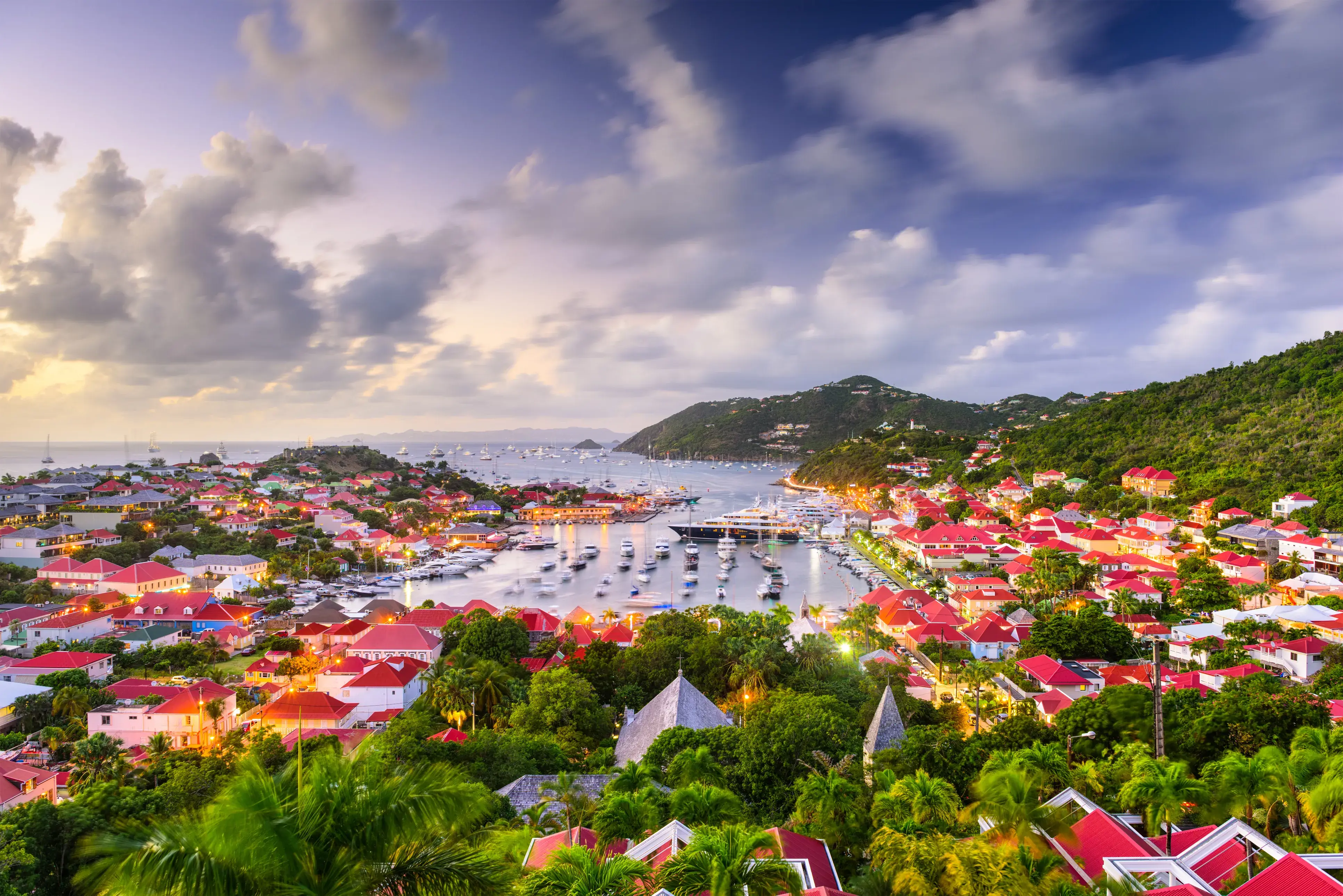 Scenic Gustavia on St Barths with red‑roofed buildings overlooking a marina filled with boats, surrounded by green hills at sunset.