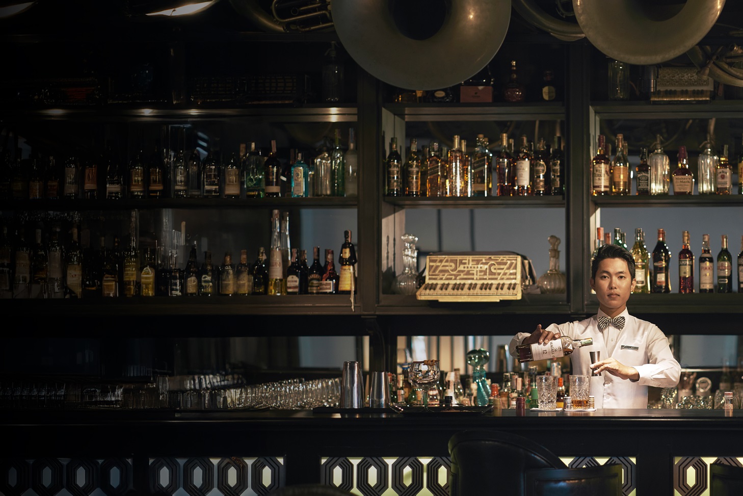 A bartender in a white shirt and bow tie prepares a drink behind a well-stocked bar with bottles and musical instruments above.