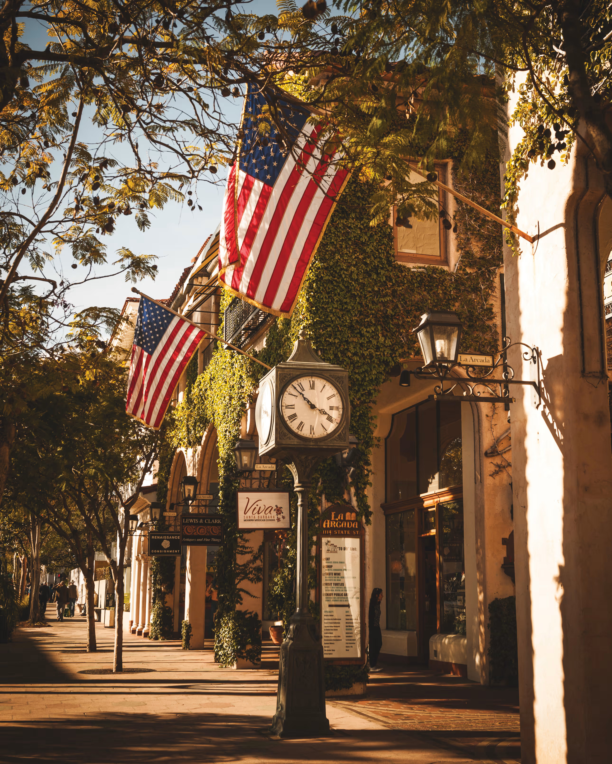 An ivy-covered building with two American flag outside and a clock tower standing on the pavement
