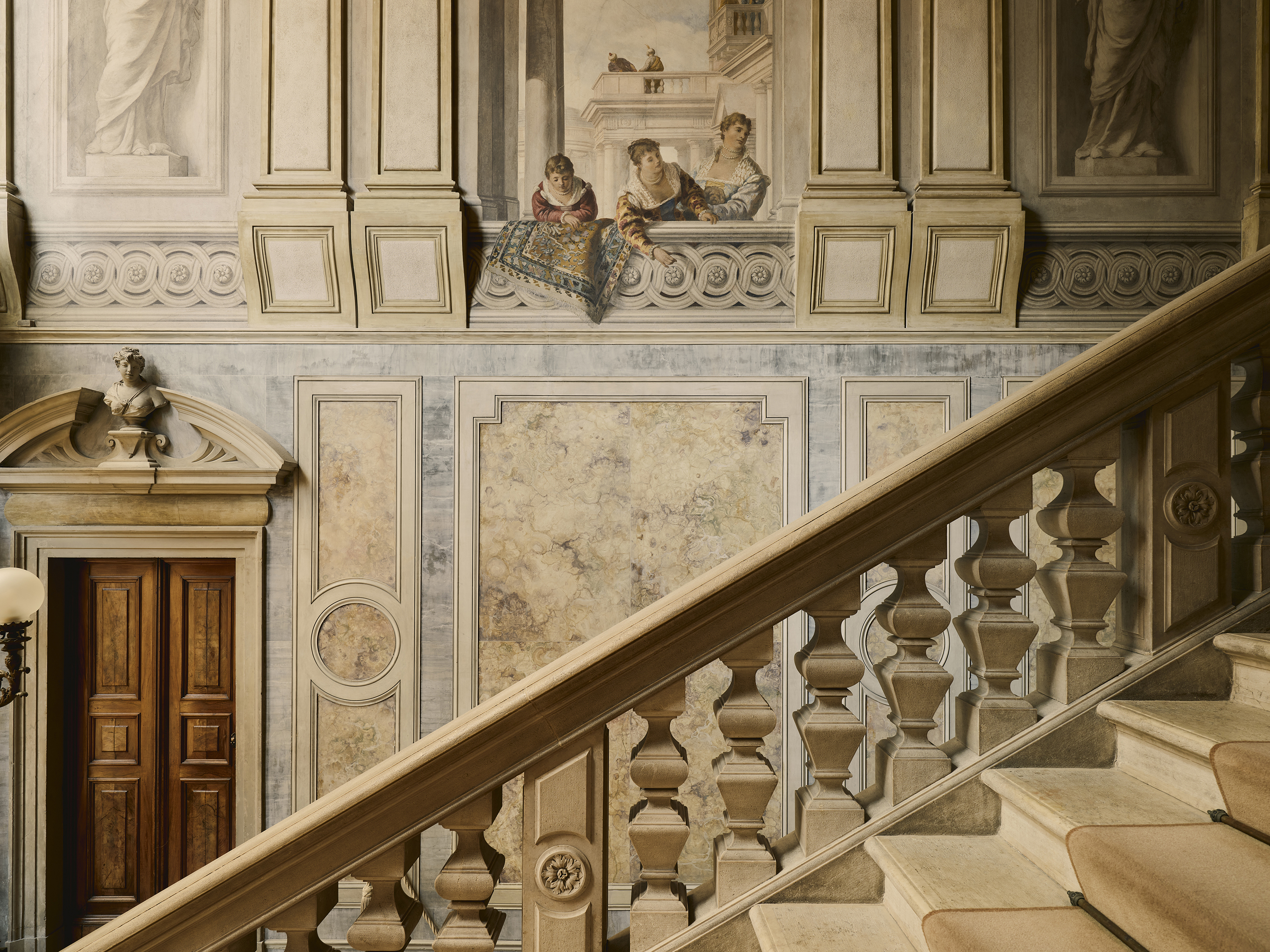 The side of a grand stone staircase opposite a painted wall featuring detailed scenes of people