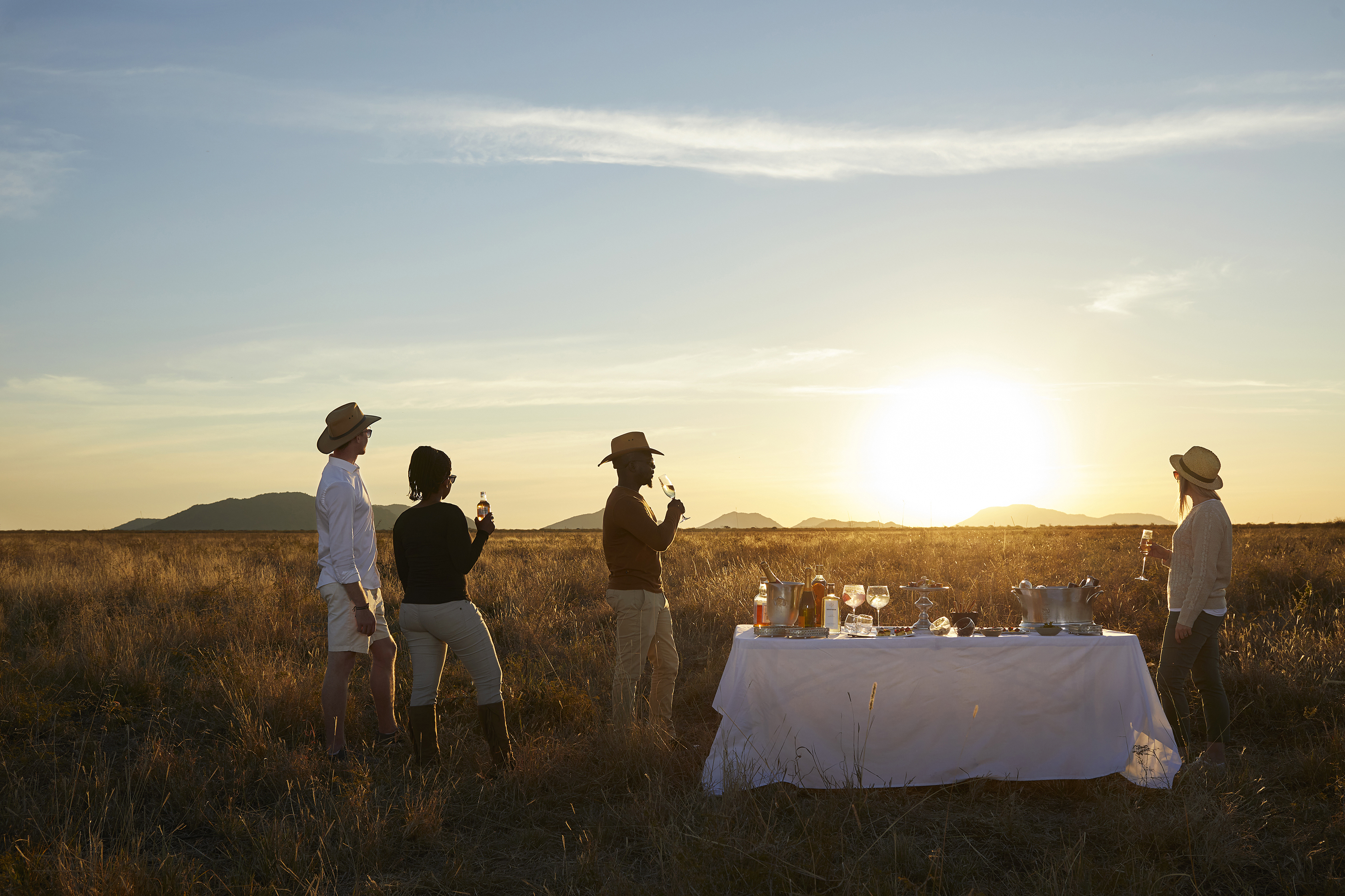 Four people enjoying sundowners from a table in the middle of the savannah as the sun sets behind