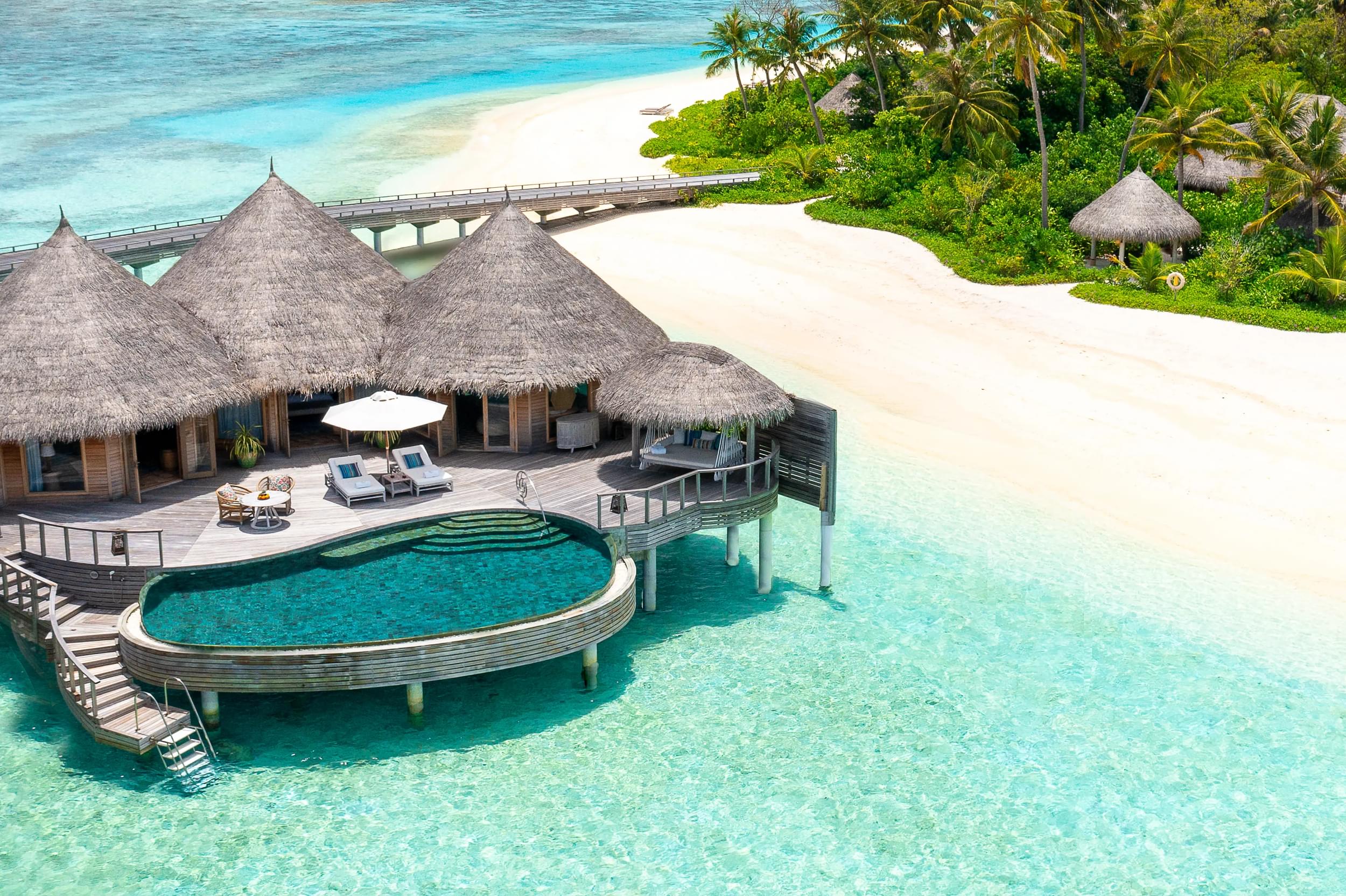 An aerial view of a luxurious overwater bungalow with a thatched roof, featuring an attached curved infinity pool, surrounded by clear turquoise waters and a white sandy beach.