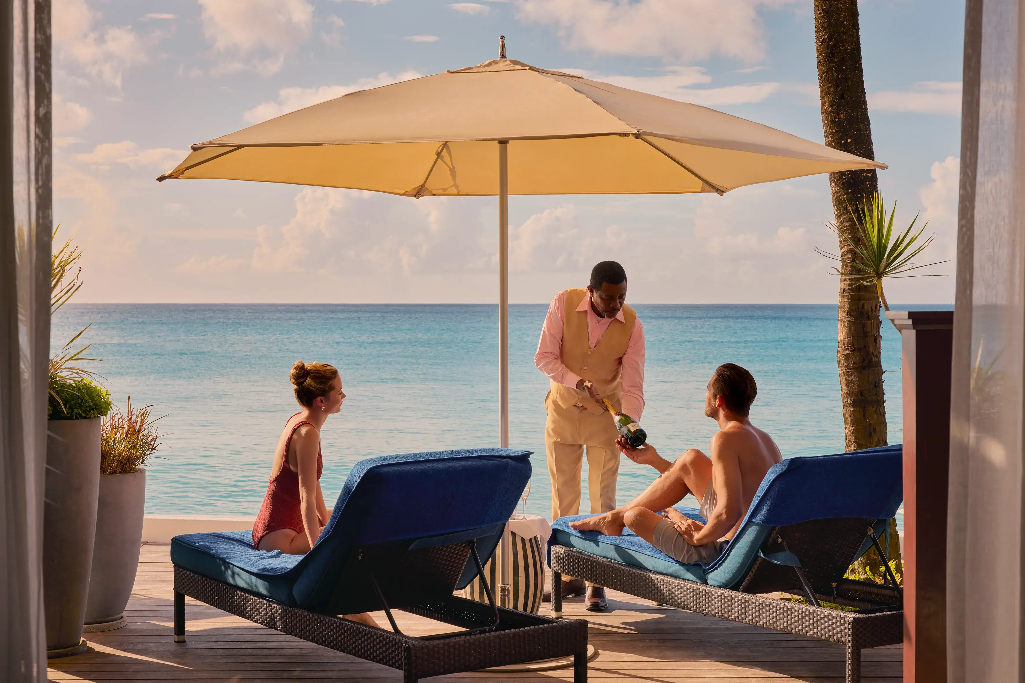 Two guests relaxing on the terrace of a Beachfront Suite at Fairmont Royal Pavilion in Barbados under a sun umbrella with a waiter displaying a bottle of champagne