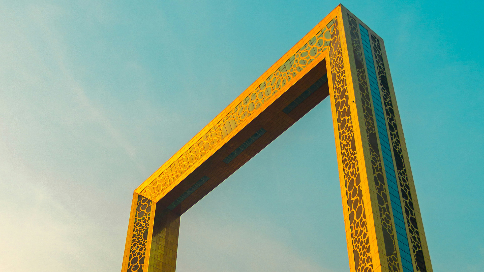 A tall, golden rectangular structure with intricate circular patterns, known as the Dubai Frame, stands against a clear blue sky, offering panoramic views of the city for families exploring modern landmarks.
