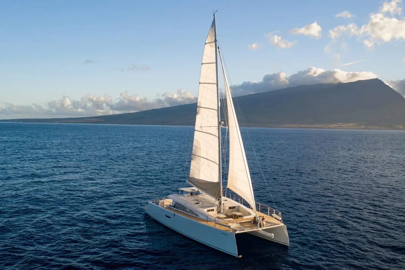 A large catamaran sailing across a calm ocean with a mountain behind under a clear sky with some fluffy clouds