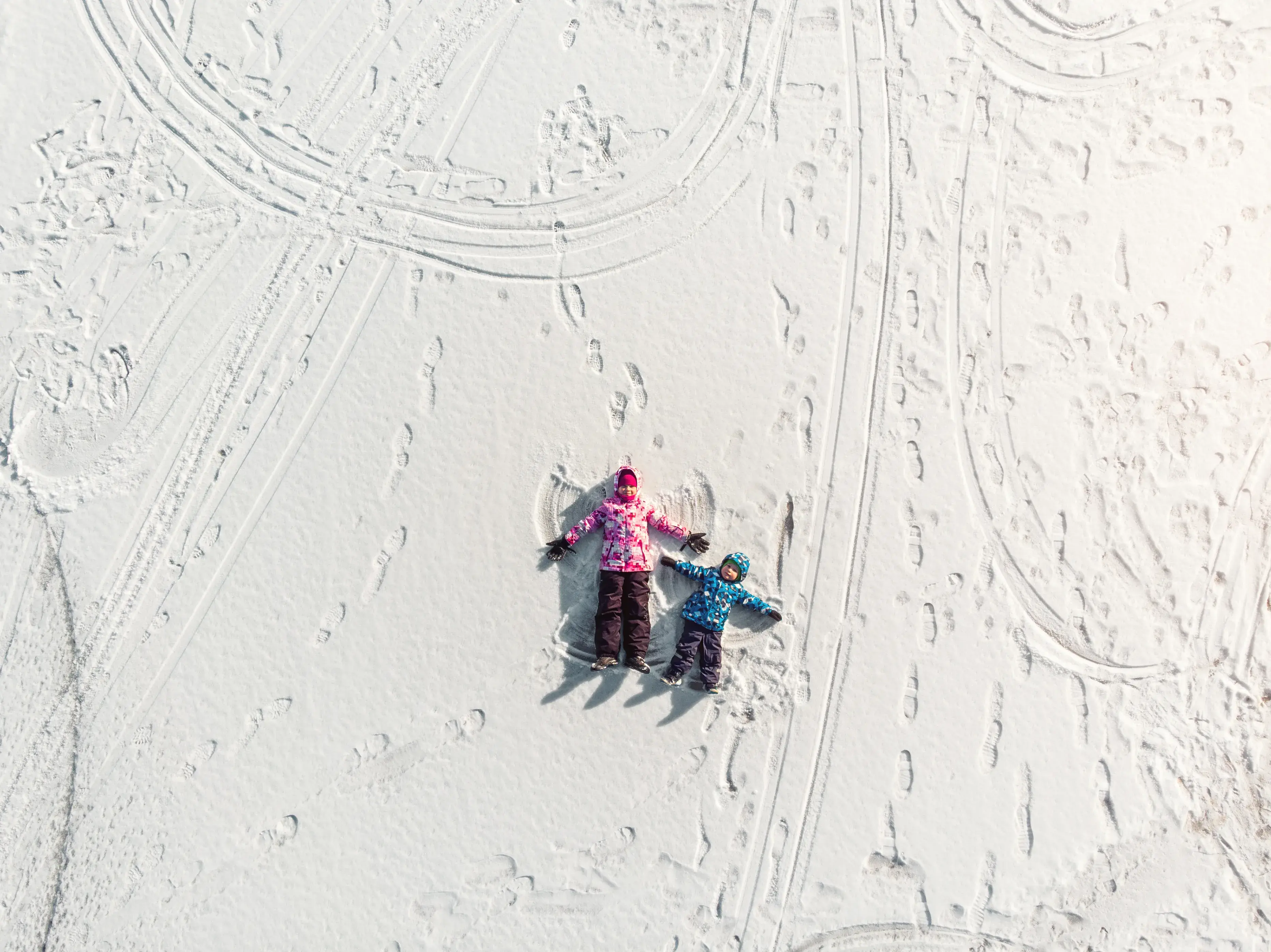 Two children lying in snow surrounded by footprints and tracks making snow angels